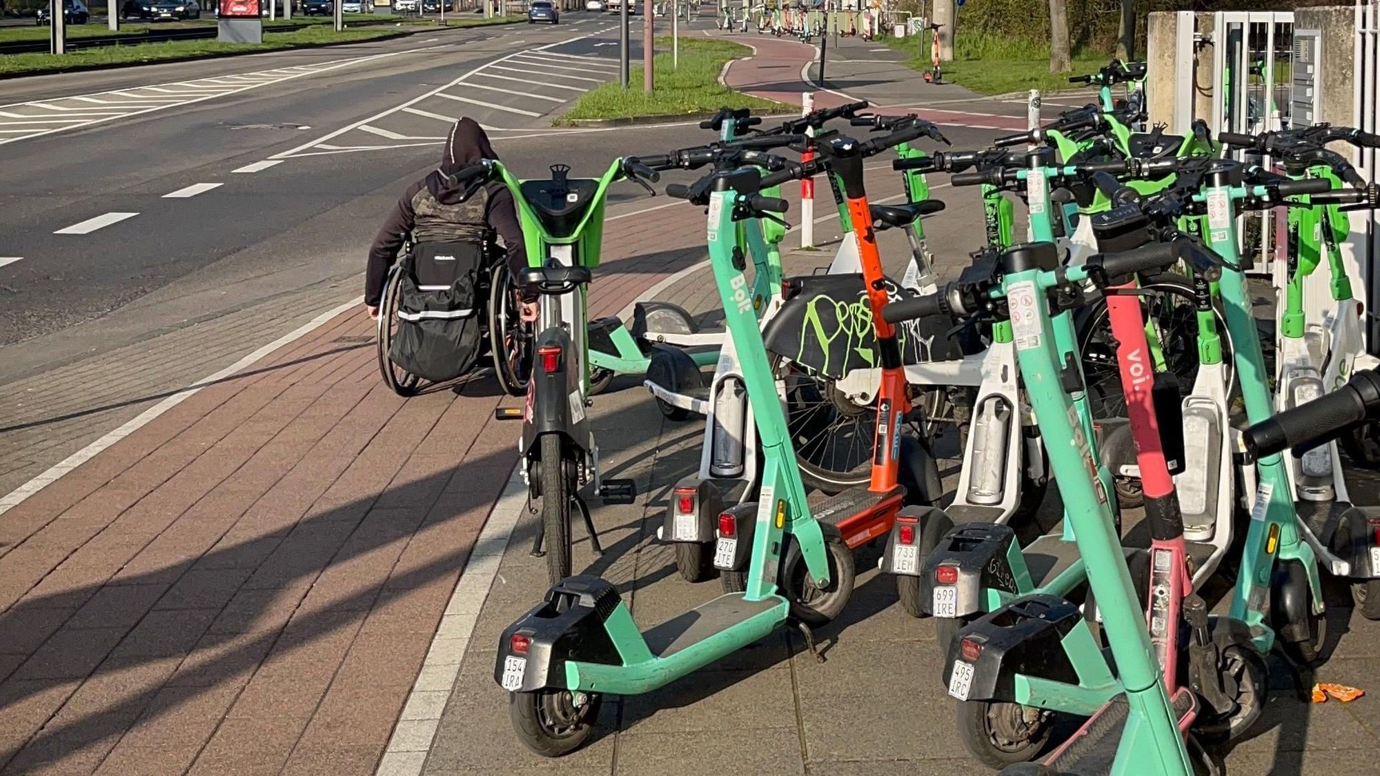 Ein Rollstuhlfahrer muss wegen der auf dem Gehweg abgestellten E-Scooter auf den Radweg ausweichen.