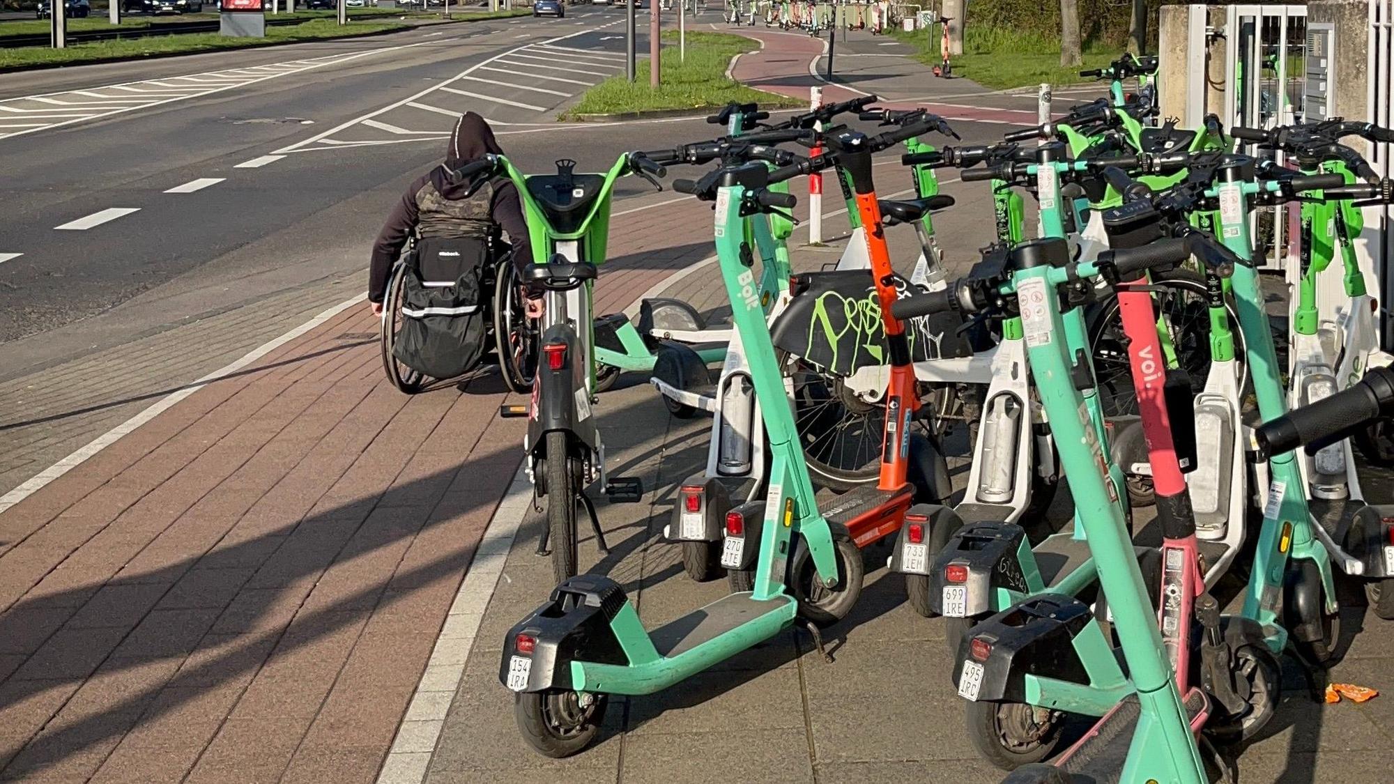 Ein Rollstuhlfahrer muss wegen der auf dem Gehweg abgestellten E-Scooter auf den Radweg ausweichen.