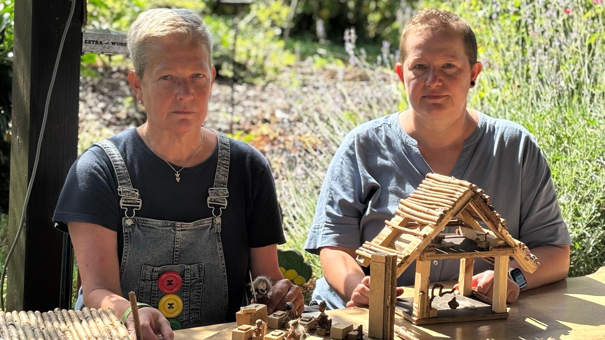Die Obengenannten sitzen mit versteinerten Gesichtern vor Teilen von Miniatur-Häusern aus Holz, die auf einem Tisch liegen.