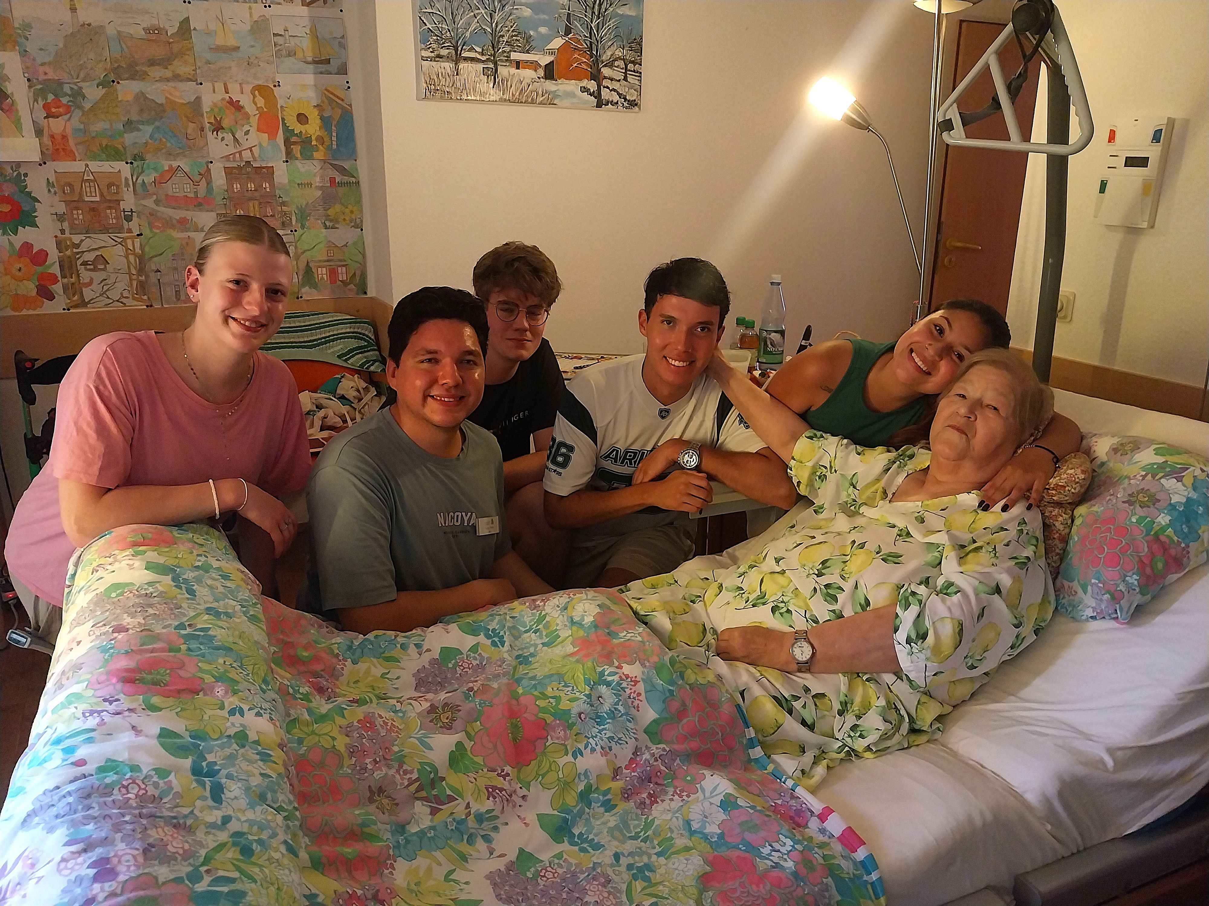 Fünf junge Menschen knien am Bett einer älteren Frau.