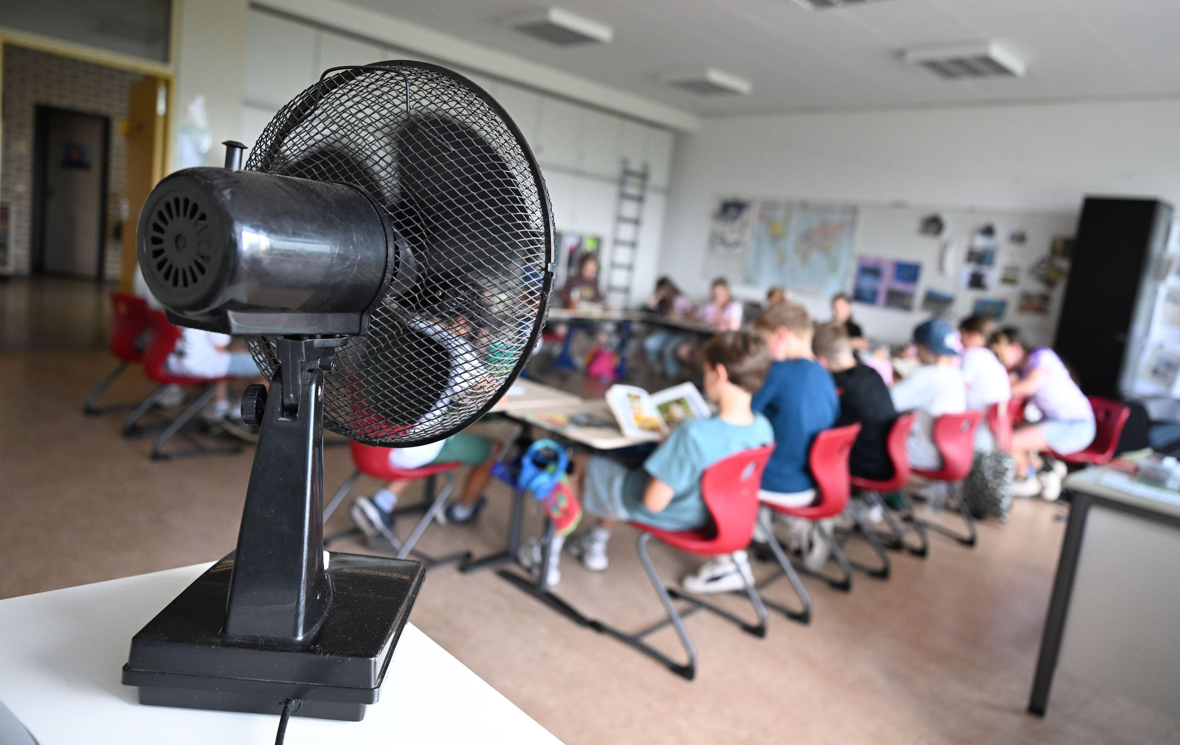 Der linke Bildrand wird dominiert von einem Ventilator, der dem im Hintergrund befindlichen Klassenraum Abkühlung verschafft. Schülerinnen und Schüler sitzen dort an Tischen.