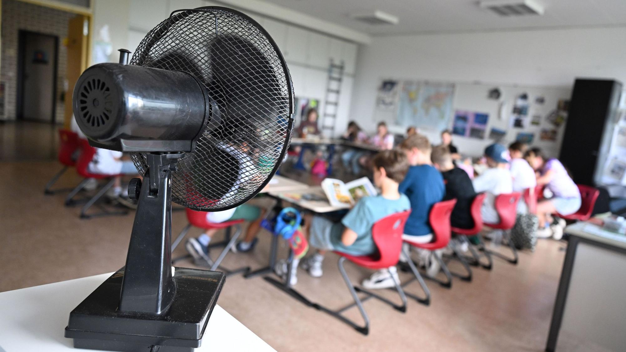 Der linke Bildrand wird dominiert von einem Ventilator, der dem im Hintergrund befindlichen Klassenraum Abkühlung verschafft. Schülerinnen und Schüler sitzen dort an Tischen.