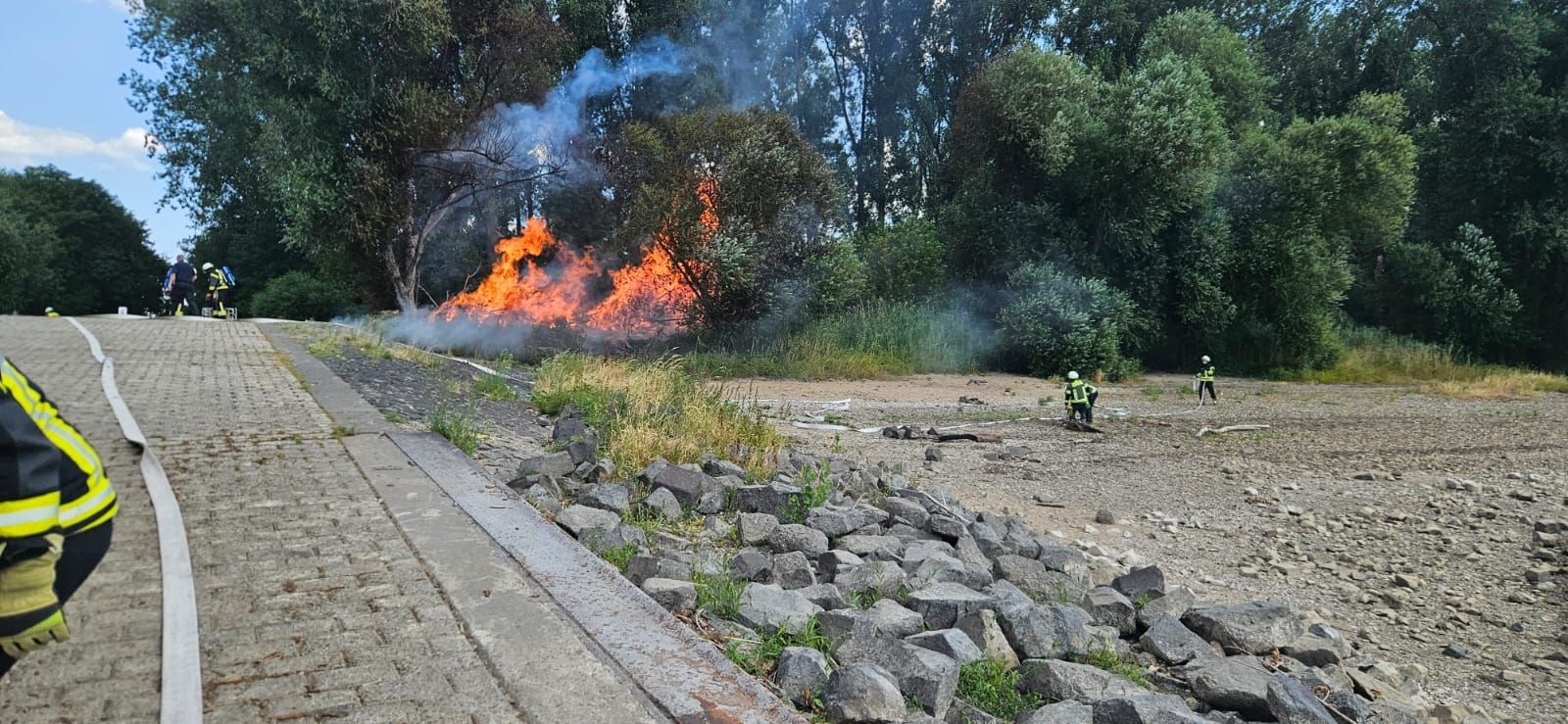 An einem Flussufer brennt es; Feuerwehrleute halten Abstand.