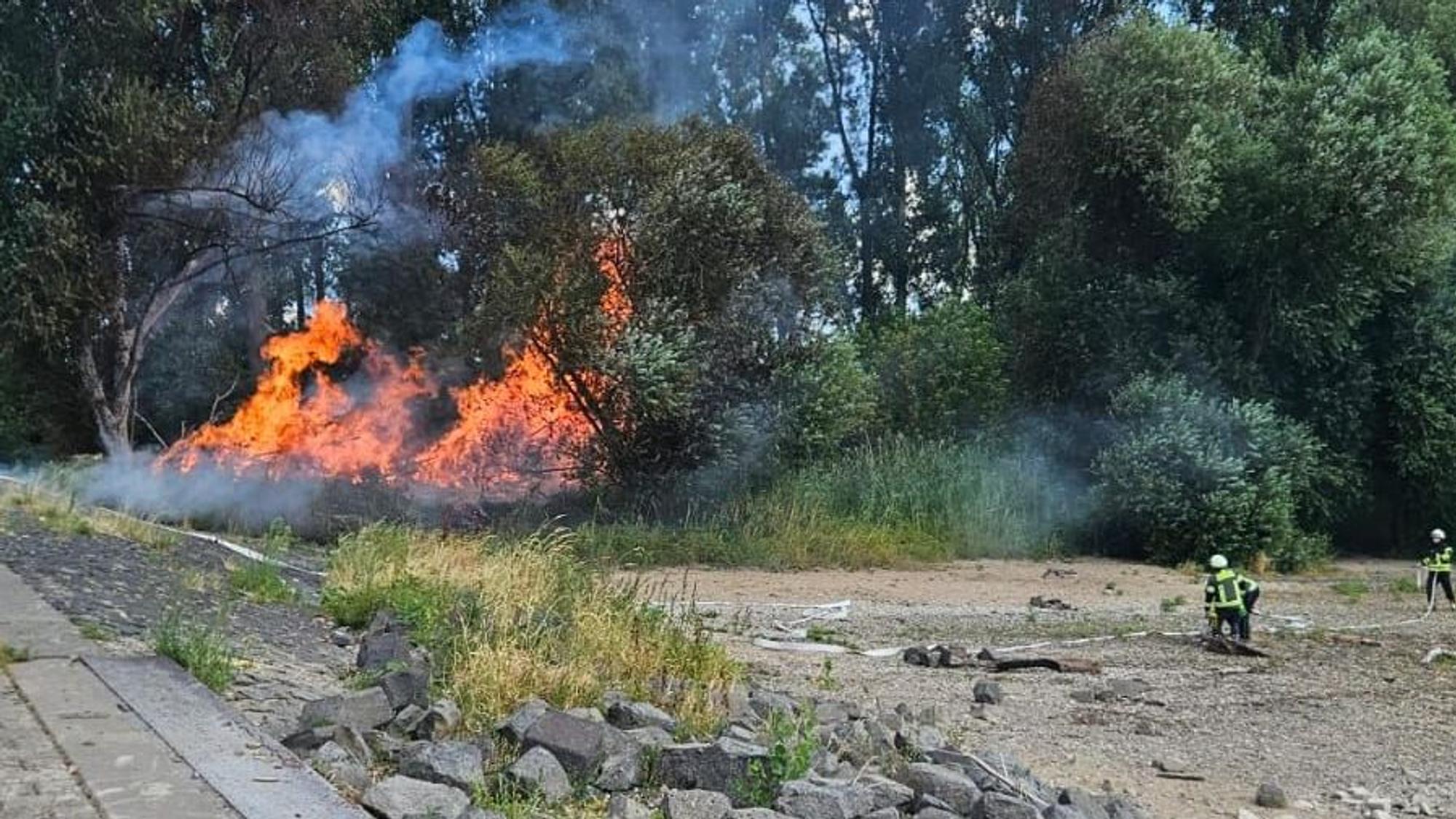 An einem Flussufer brennt es; Feuerwehrleute halten Abstand.