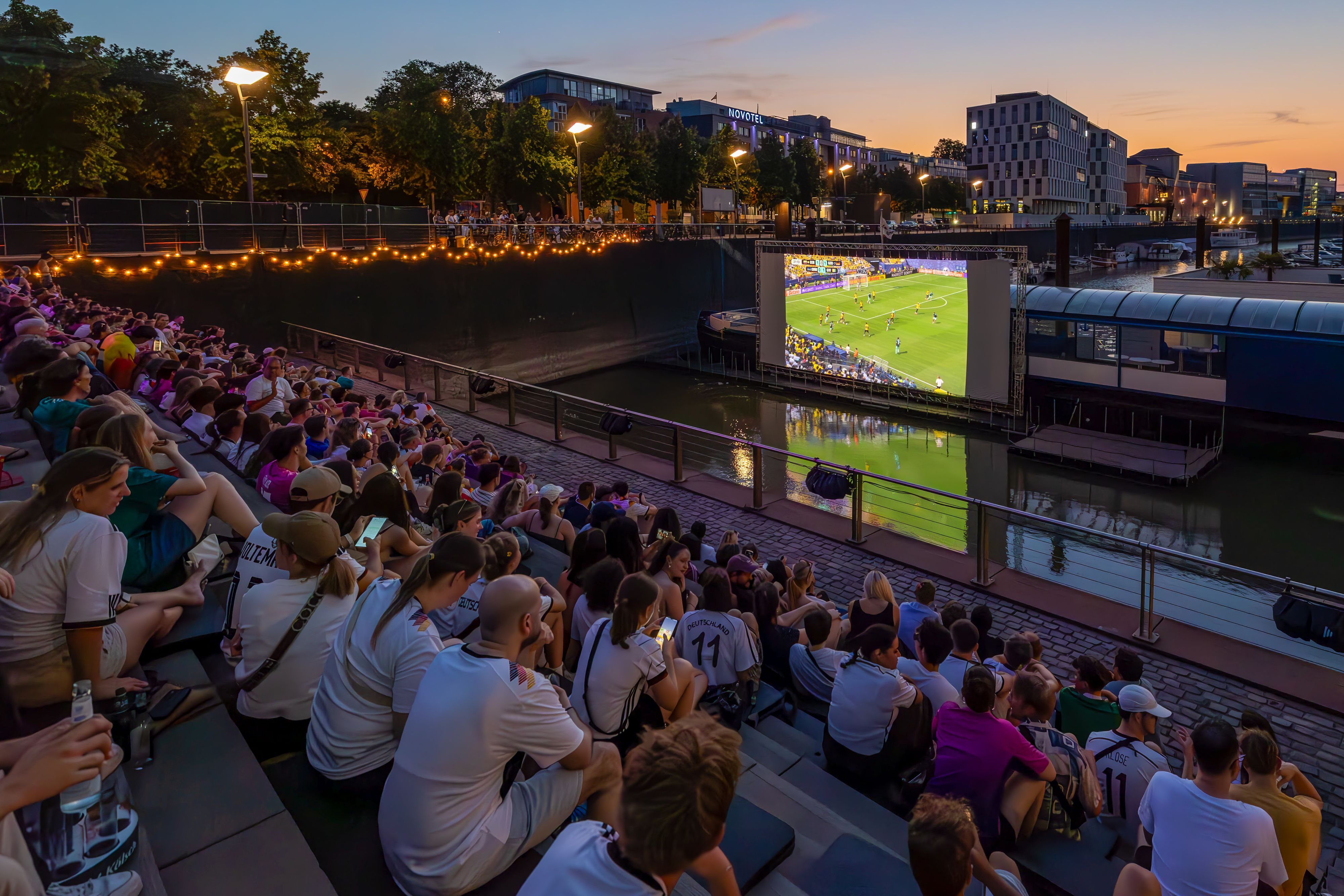 Zuschauende im Open-Air-Kino gucken die Fußball-Weltmeisterschaft, es läuft ein Spiel mit deutscher Beteiligung.