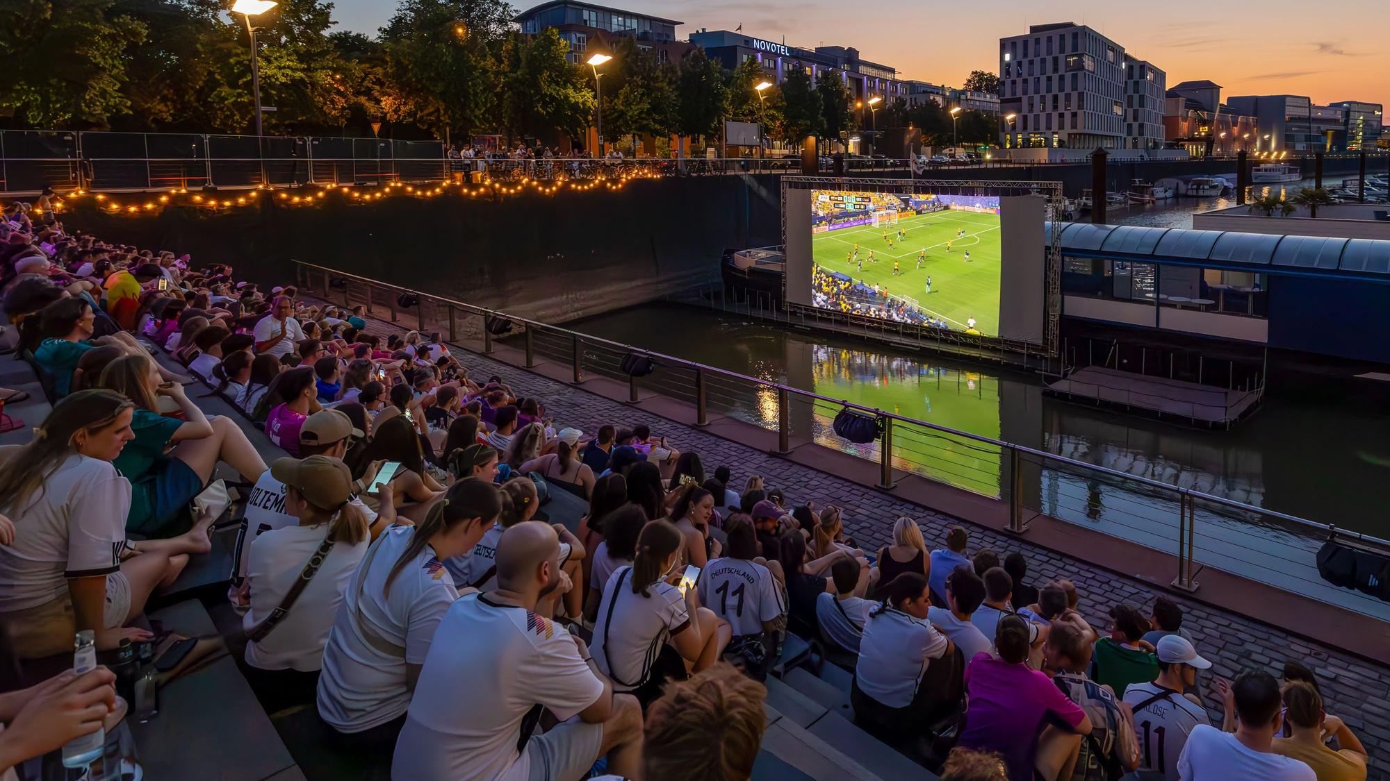 Zuschauende im Open-Air-Kino gucken die Fußball-Weltmeisterschaft, es läuft ein Spiel mit deutscher Beteiligung.