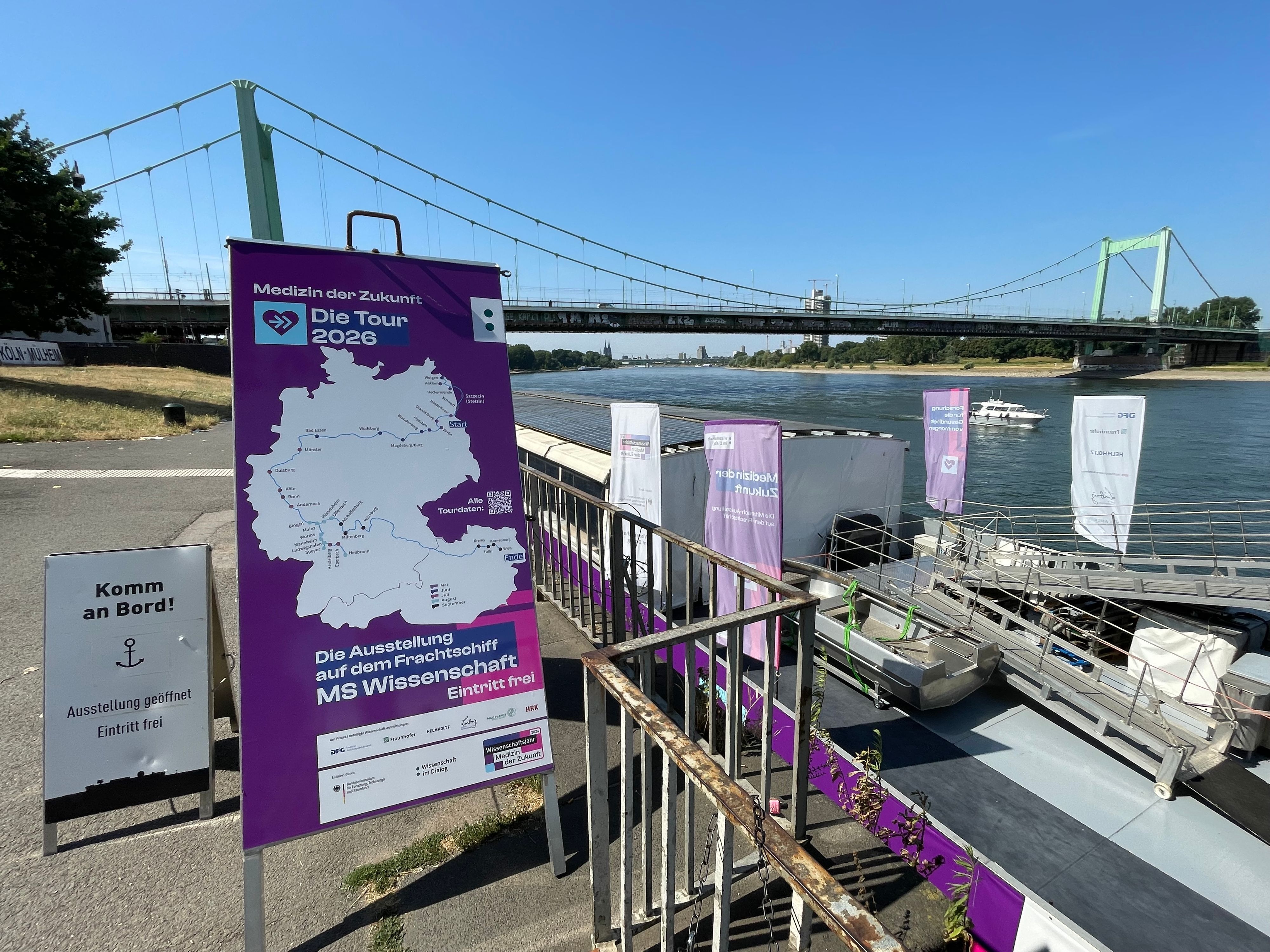 Ein lila Plakat, das die Stationen des Schiffes zeigt, das dahinter am Ufer liegt. Im Hintergrund der Rhein und die Mülheimer Brücke in Köln.