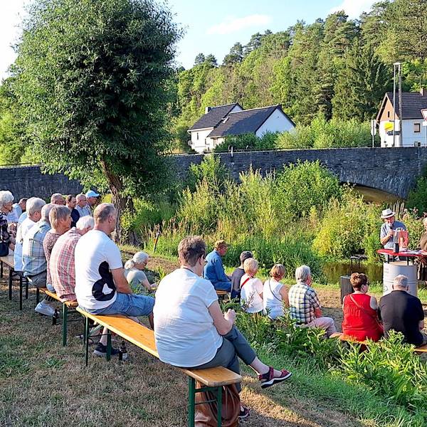 Menschen sitzen auf Holzbänken am Flussufer. Sie richten den Blick auf einen Redner, der an einem Pult steht.