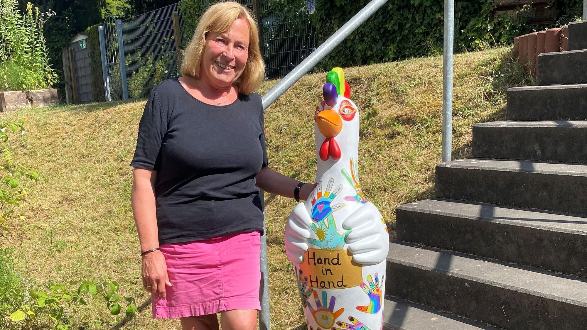 Eine Frau steht neben einer bunt bemalten Skulptur, einem Huhn