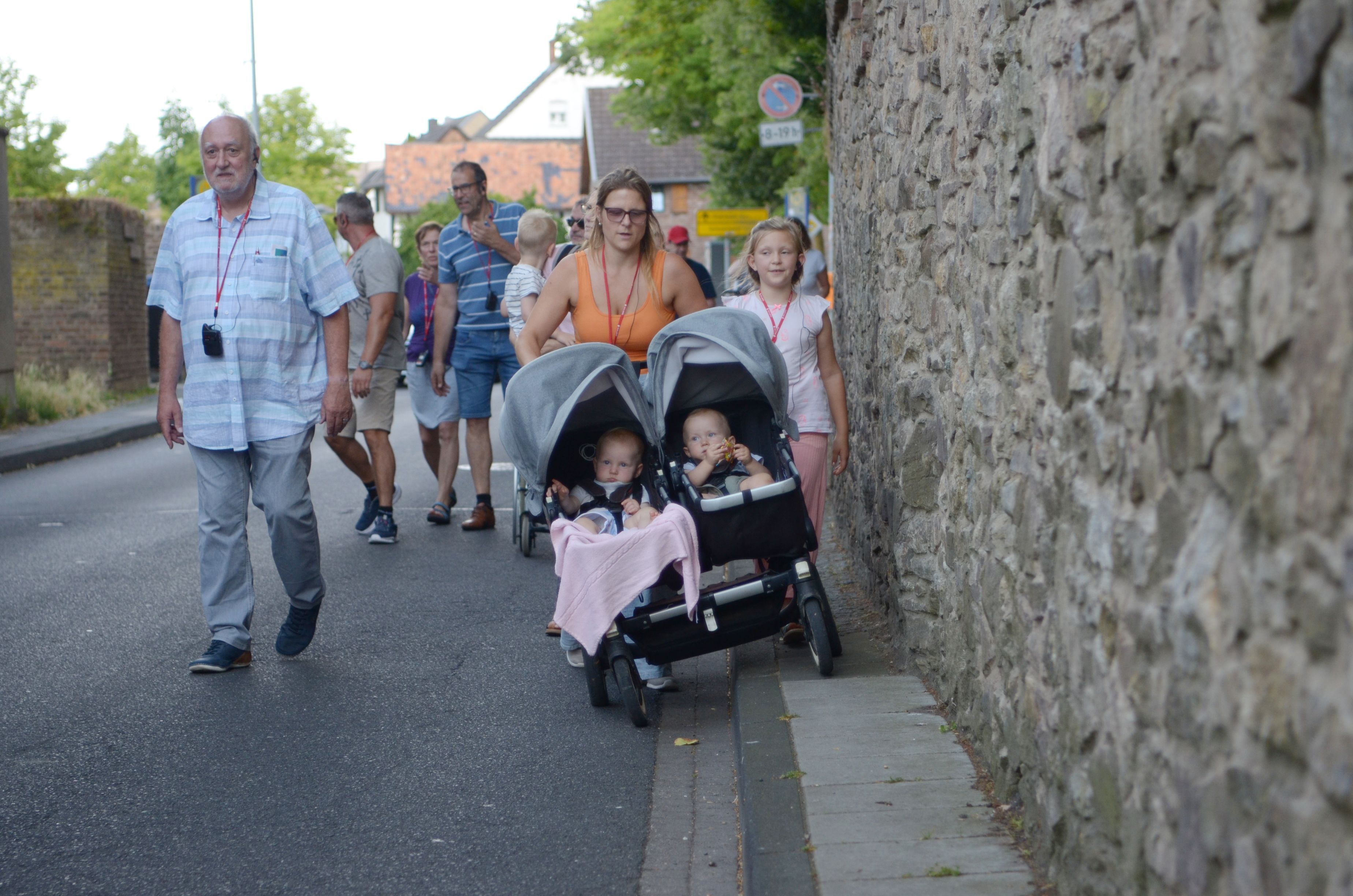 In einer Gruppe von Menschen geht eine Frau, die mit einer Seite ihres Kinderwagens auf die Fahrbahn ausweichen muss, weil der Gehweg zu schmal ist.