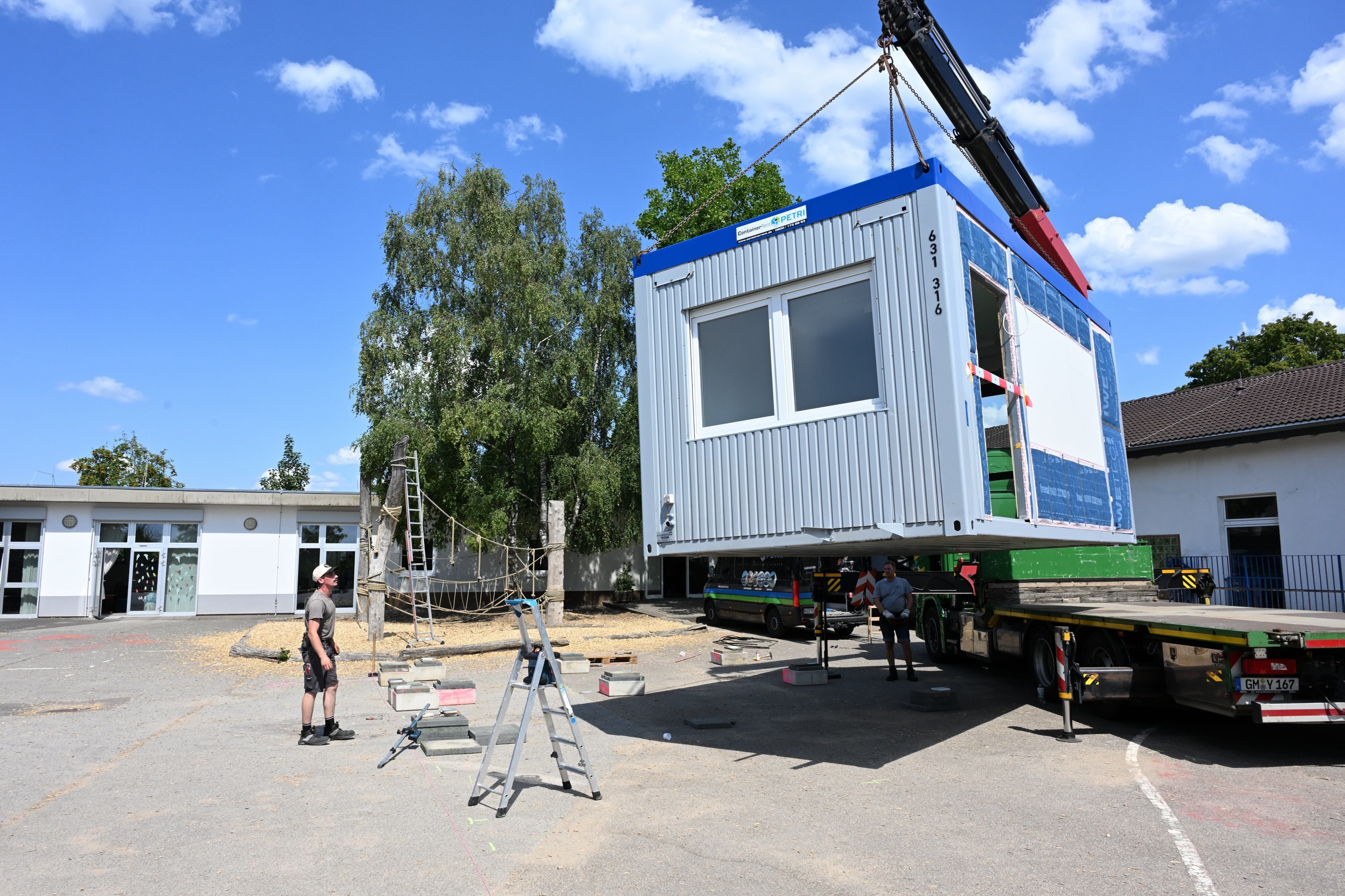 Ein Container hängt am Haken eines Krans und wird auf einen Schulhof herabgelassen.