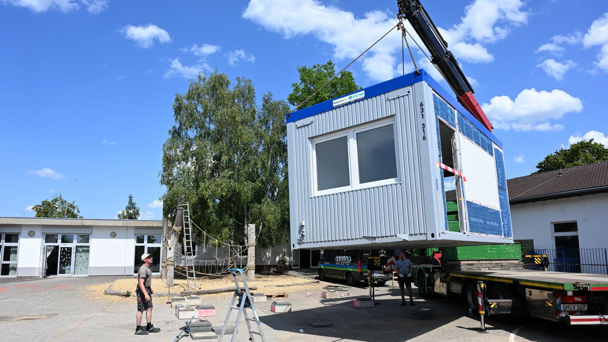Ein Container hängt am Haken eines Krans und wird auf einen Schulhof herabgelassen.