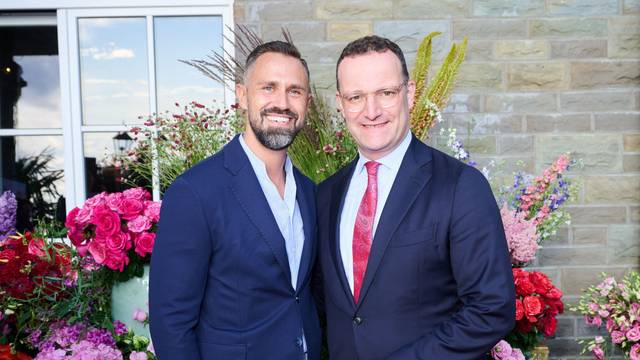 Jens Spahn und sein Ehemann Daniel Funke stehen vor einer mit Blumen geschmückten Hauswand und lächeln in die Kamera. Beide tragen blaue Anzugjacken.