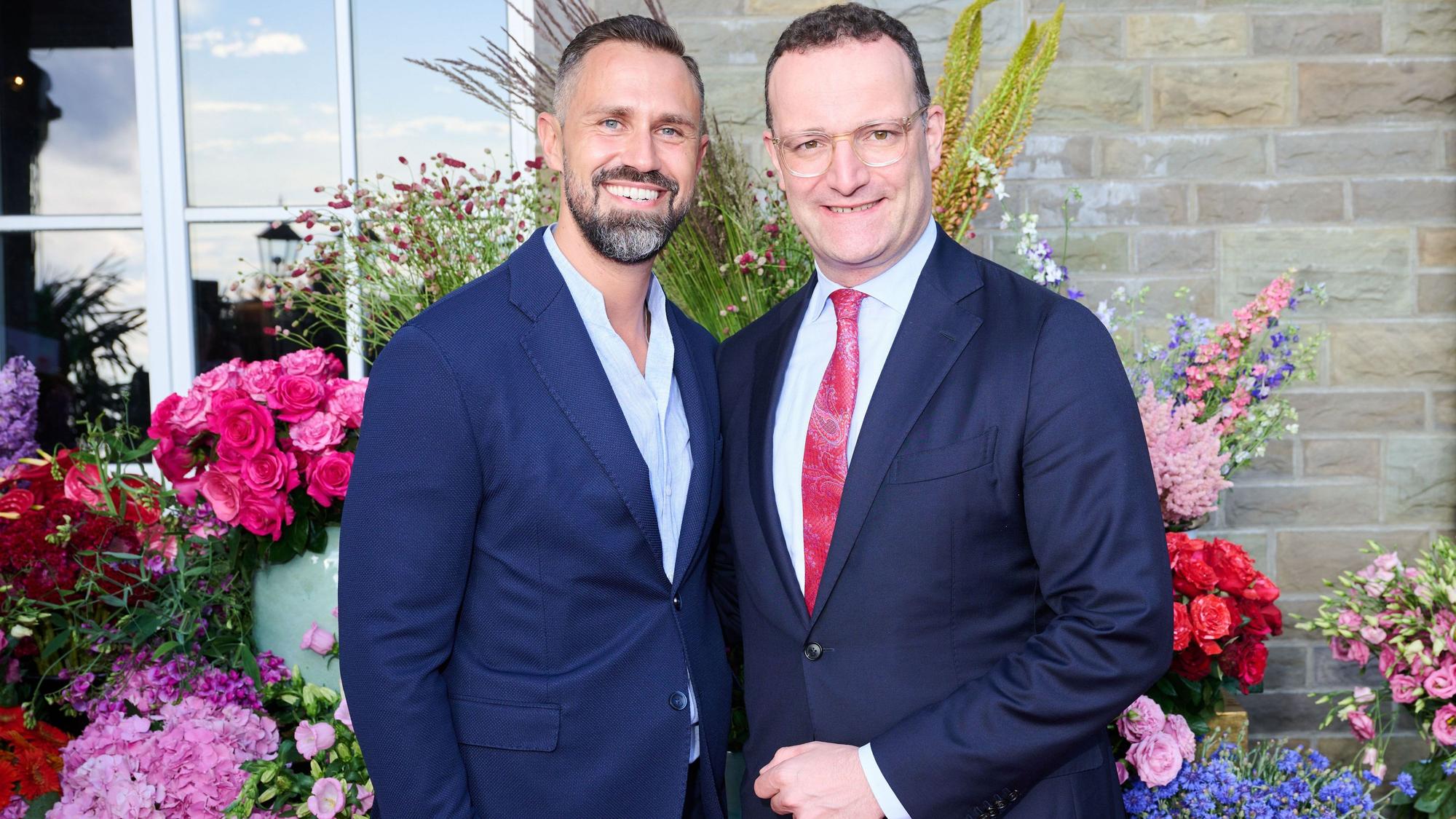 Jens Spahn und sein Ehemann Daniel Funke stehen vor einer mit Blumen geschmückten Hauswand und lächeln in die Kamera. Beide tragen blaue Anzugjacken.