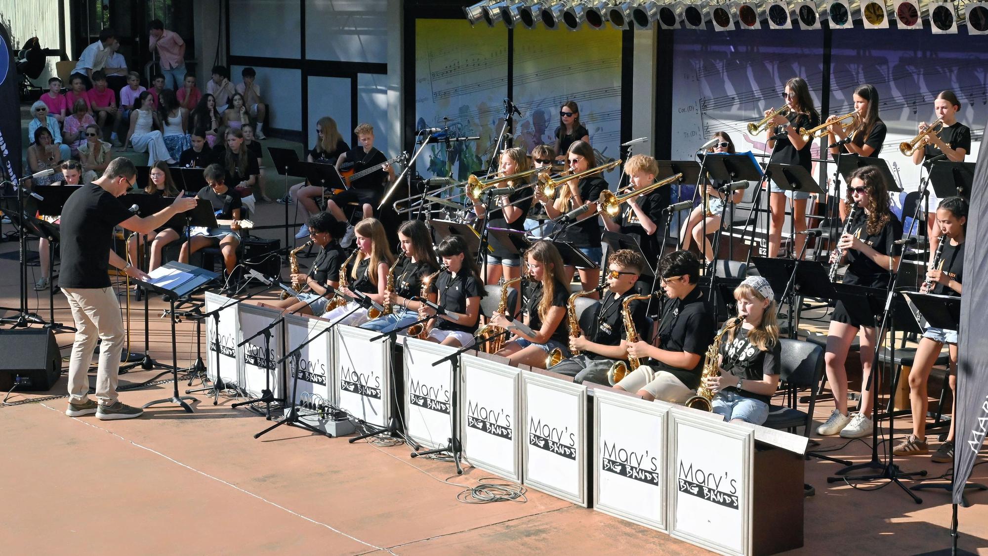 Die Musikerinnen und Musiker spielen teils sitzend, teils stehend. Davor steht der Bandleader.