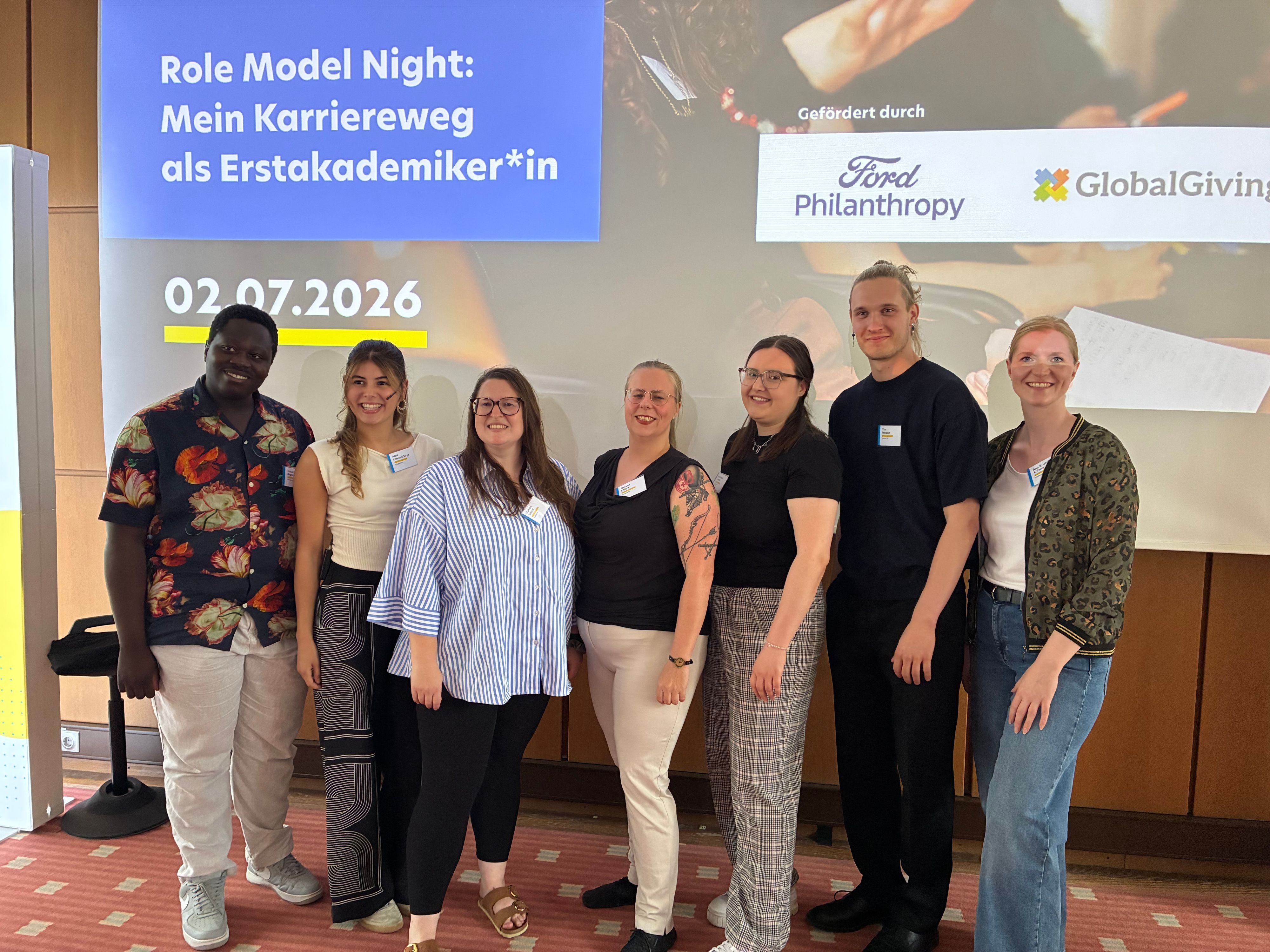 Sieben Personen stehen vor einer Leinwand, auf der steht: „Role Model Night: Mein Karriereweg als Erstakademiker*in“, darunter das Datum (02.07.2026) sowie daneben „Gefördert durch Ford Philanthropy und Global Giving“.