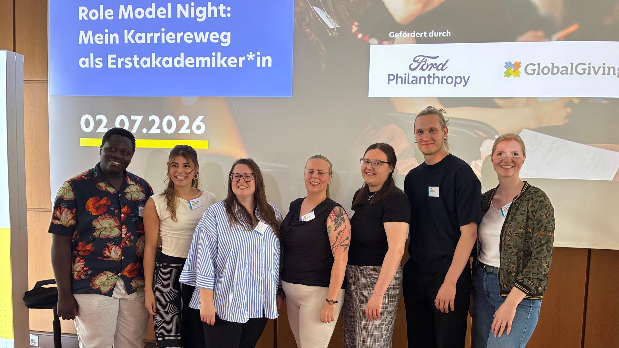 Sieben Personen stehen vor einer Leinwand, auf der steht: „Role Model Night: Mein Karriereweg als Erstakademiker*in“, darunter das Datum (02.07.2026) sowie daneben „Gefördert durch Ford Philanthropy und Global Giving“.