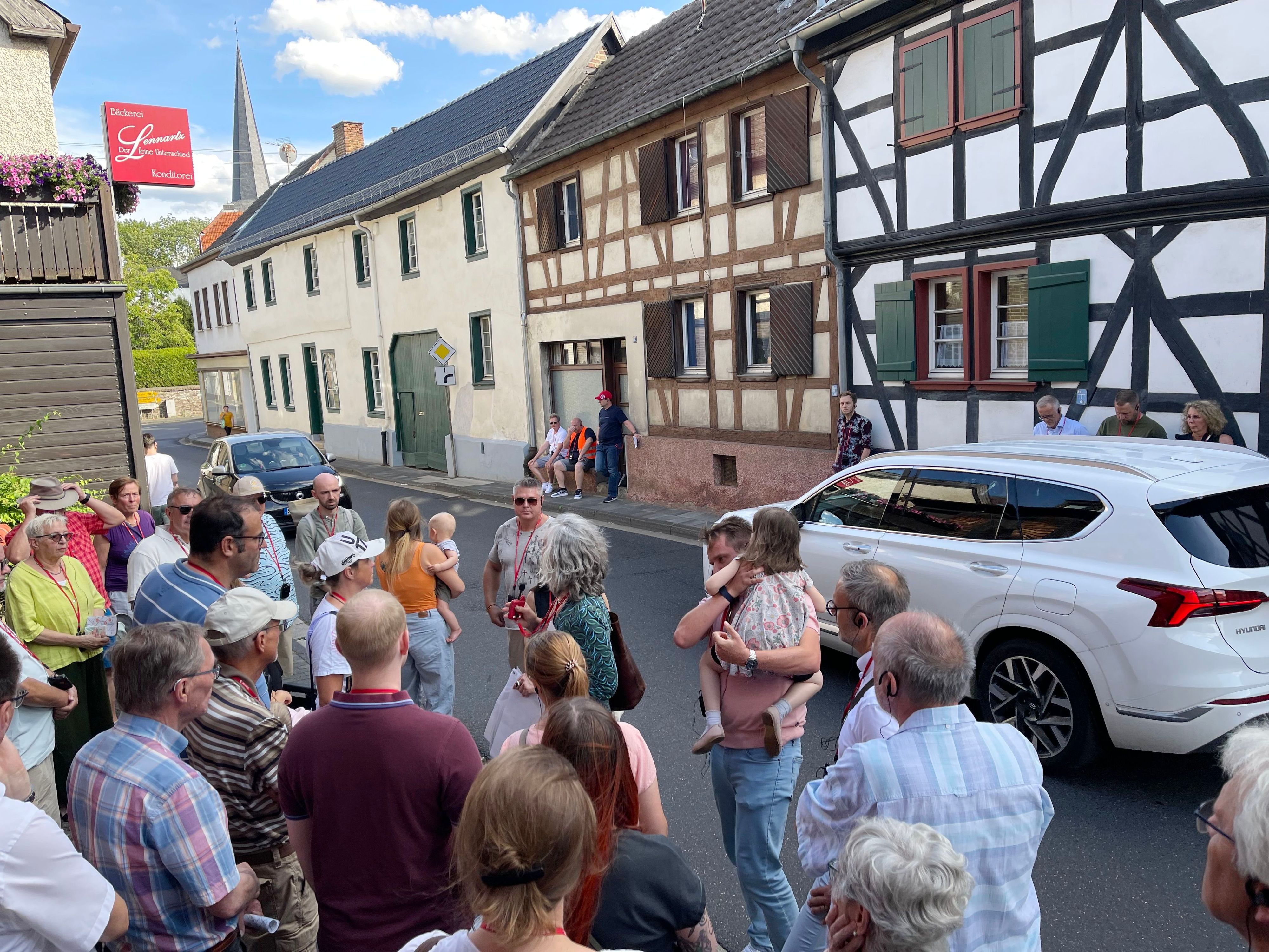Eine Gruppe von Menschen steht am Straßenrand, Autos fahren an ihnen vorbei.
