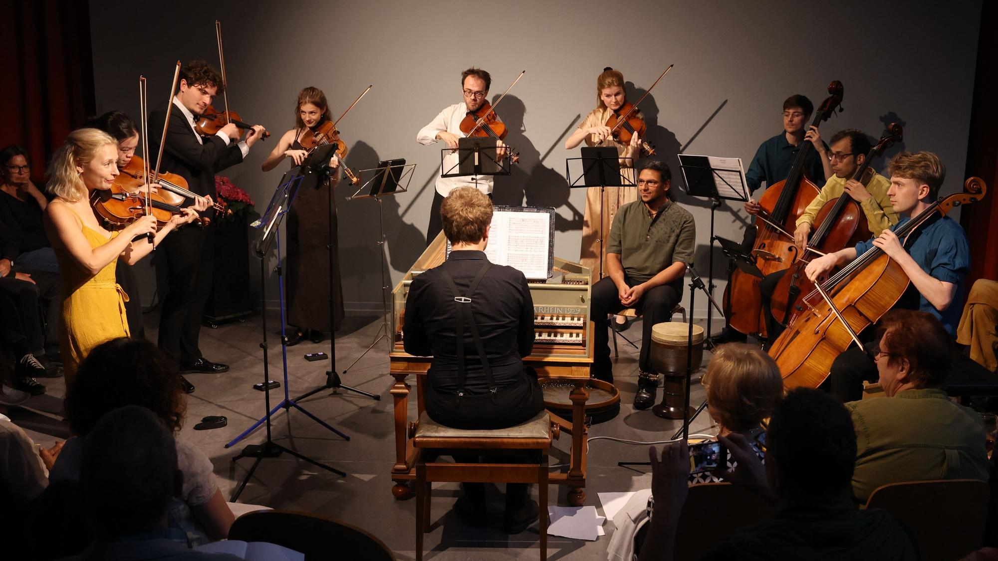 Neun Streichmusiker und ein Mann am Klavier spielen bei einem Konzert in einer ehemaligen Klosterkapelle vor Publikum.