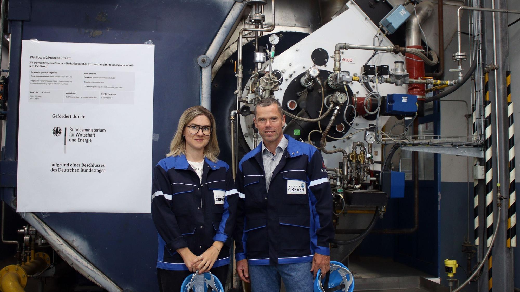 Eine Frau und ein Mann, beide halten in den Händen einen Helm, stehen vor einem Dampfkessel.