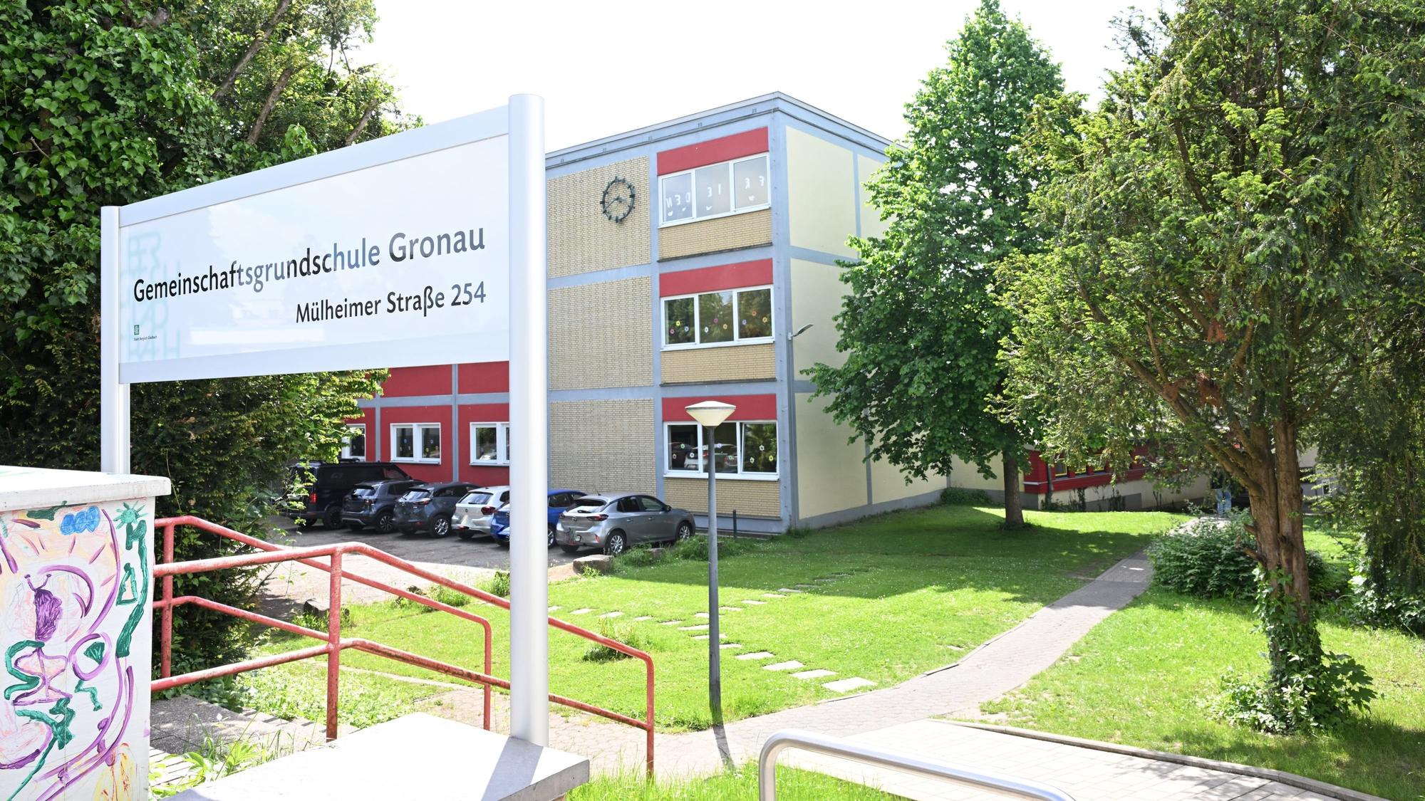 Gemeinschaftsgrundschule Gronau steht auf einem Schild im Vordergrund. Im Hintergrund ein Flachdach-Schulgebäude