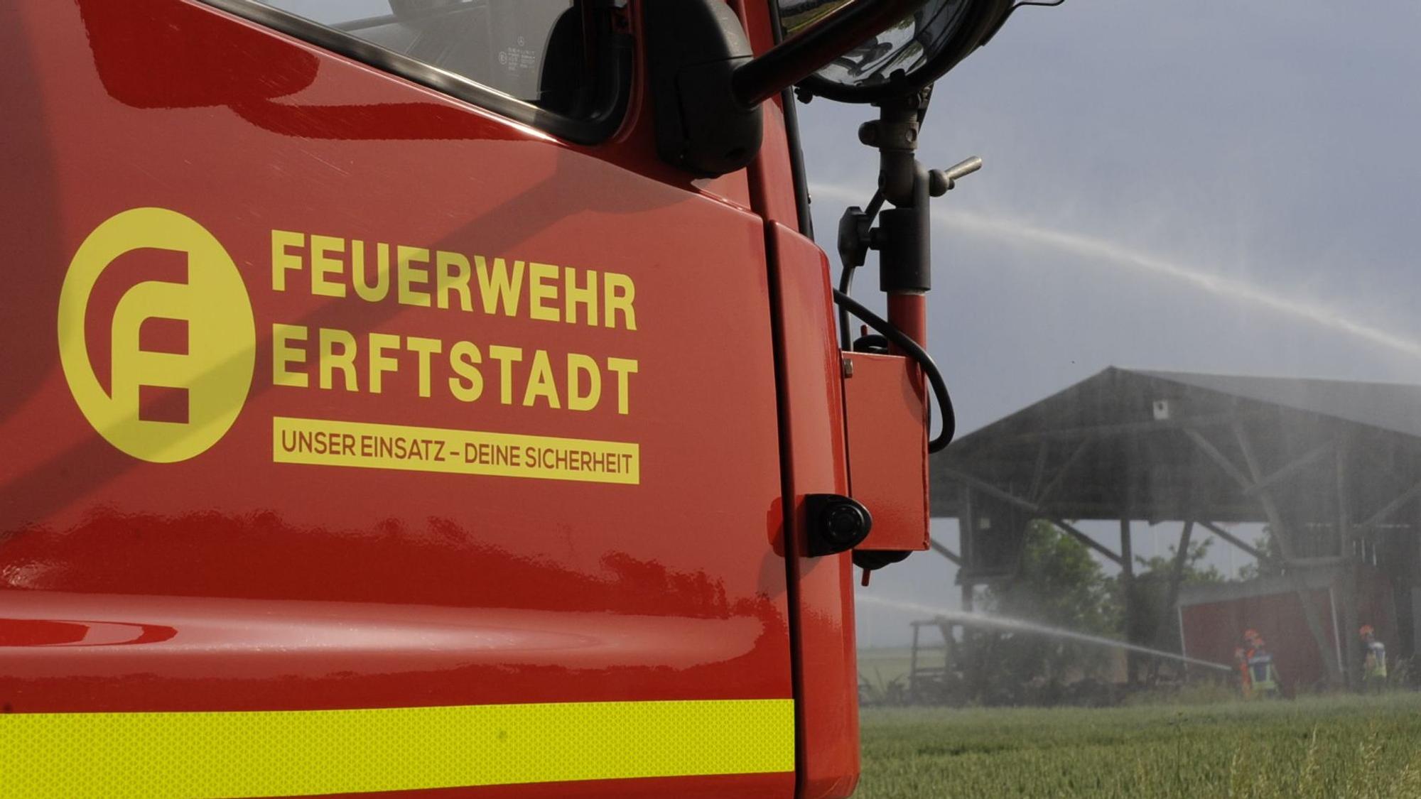 Ein Einsatzfahrzeug der Feuerwehr Erftstadt steht an einem Feld, dahinter sind eine Scheune und Löscharbeiten zu sehen.