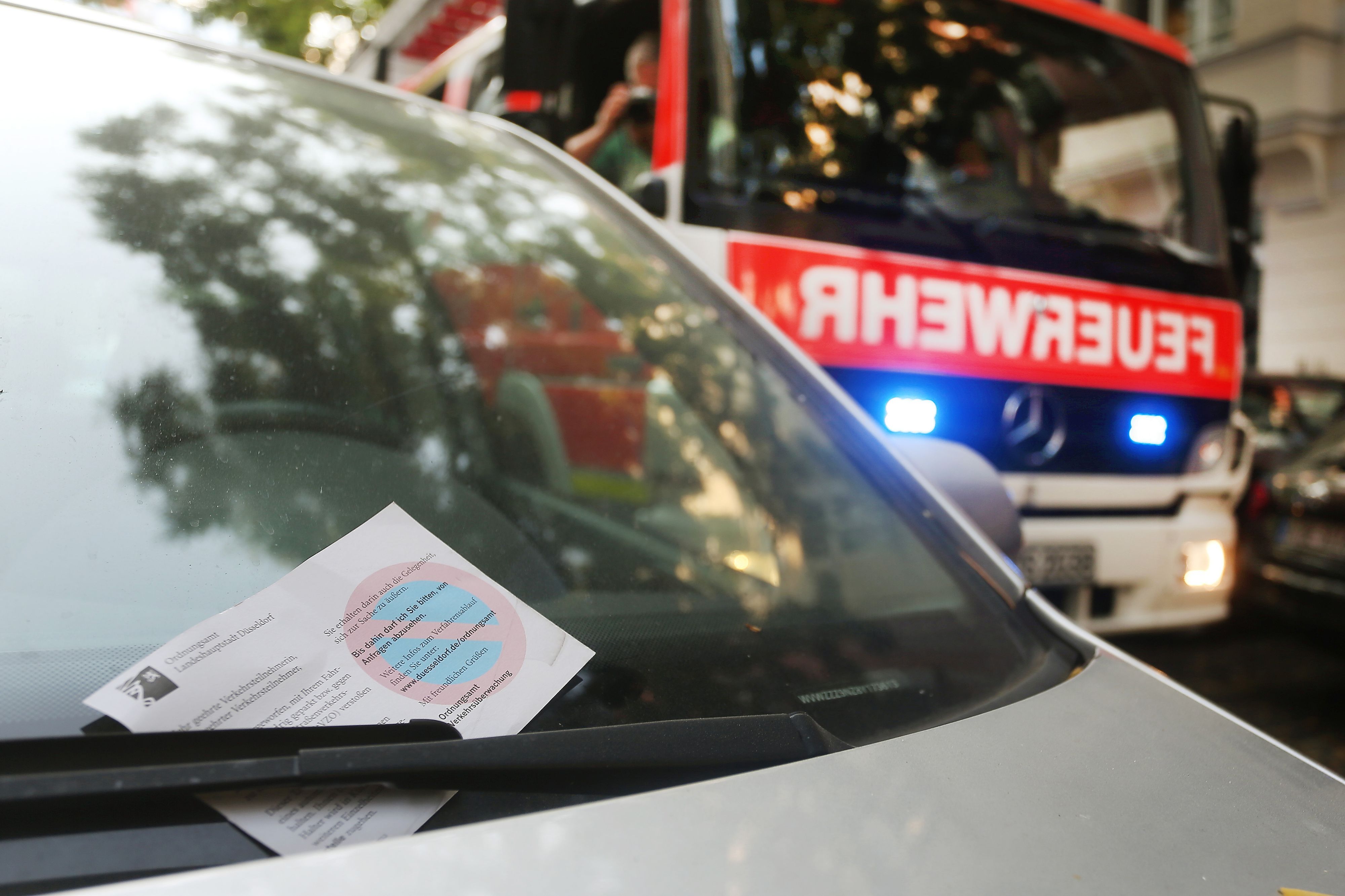 Ein falsch geparktes Auto mit einem Knöllchen, im Hintergrund ist ein Feuerwehrauto mit Blaulicht zu sehen. (Symbolbild)