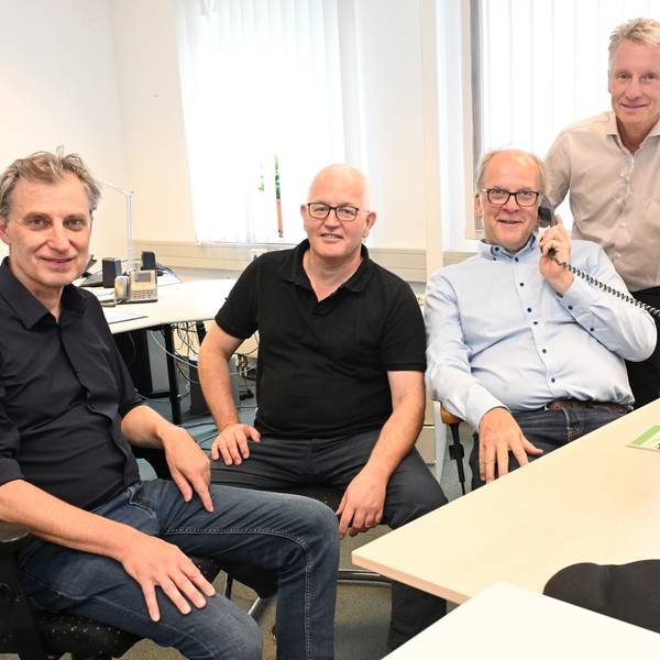 Drei Männer sitzen, einer von ihnen hält einen Telefonhörer in der Hand, ein vierter steht neben ihnen