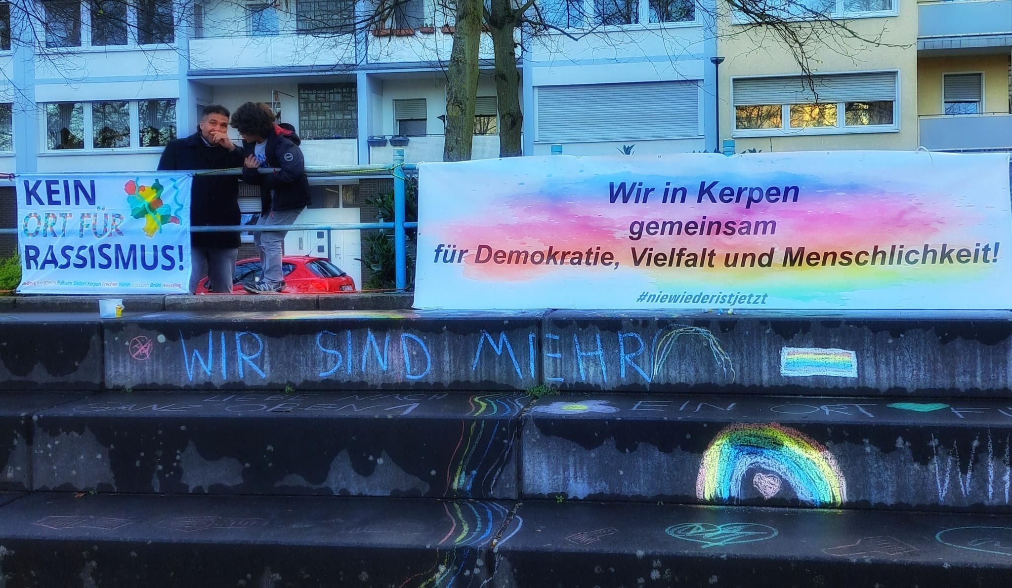 Das Foto zeigt ein Plakat, daneben zwei Menschen.