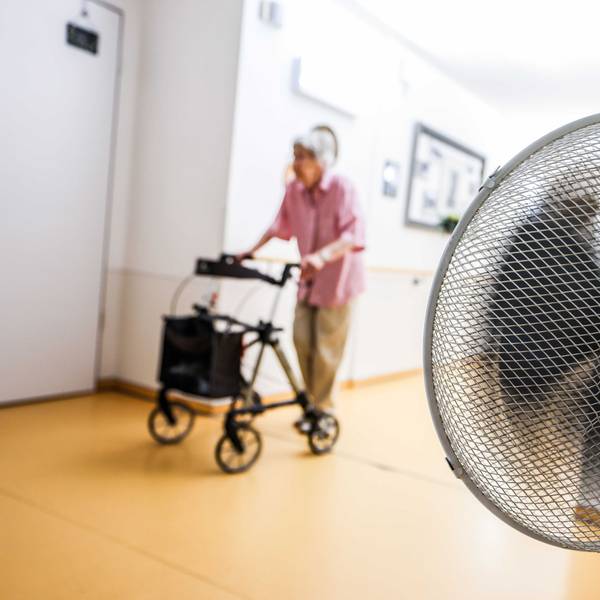 Eine Seniorin schiebt ihren Rollator über einen Flur. Rechts ist im Vordergrund ein Ventilator zu sehen.