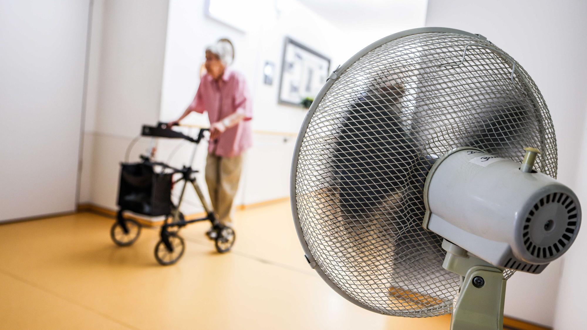 Eine Seniorin schiebt ihren Rollator über einen Flur. Rechts ist im Vordergrund ein Ventilator zu sehen.