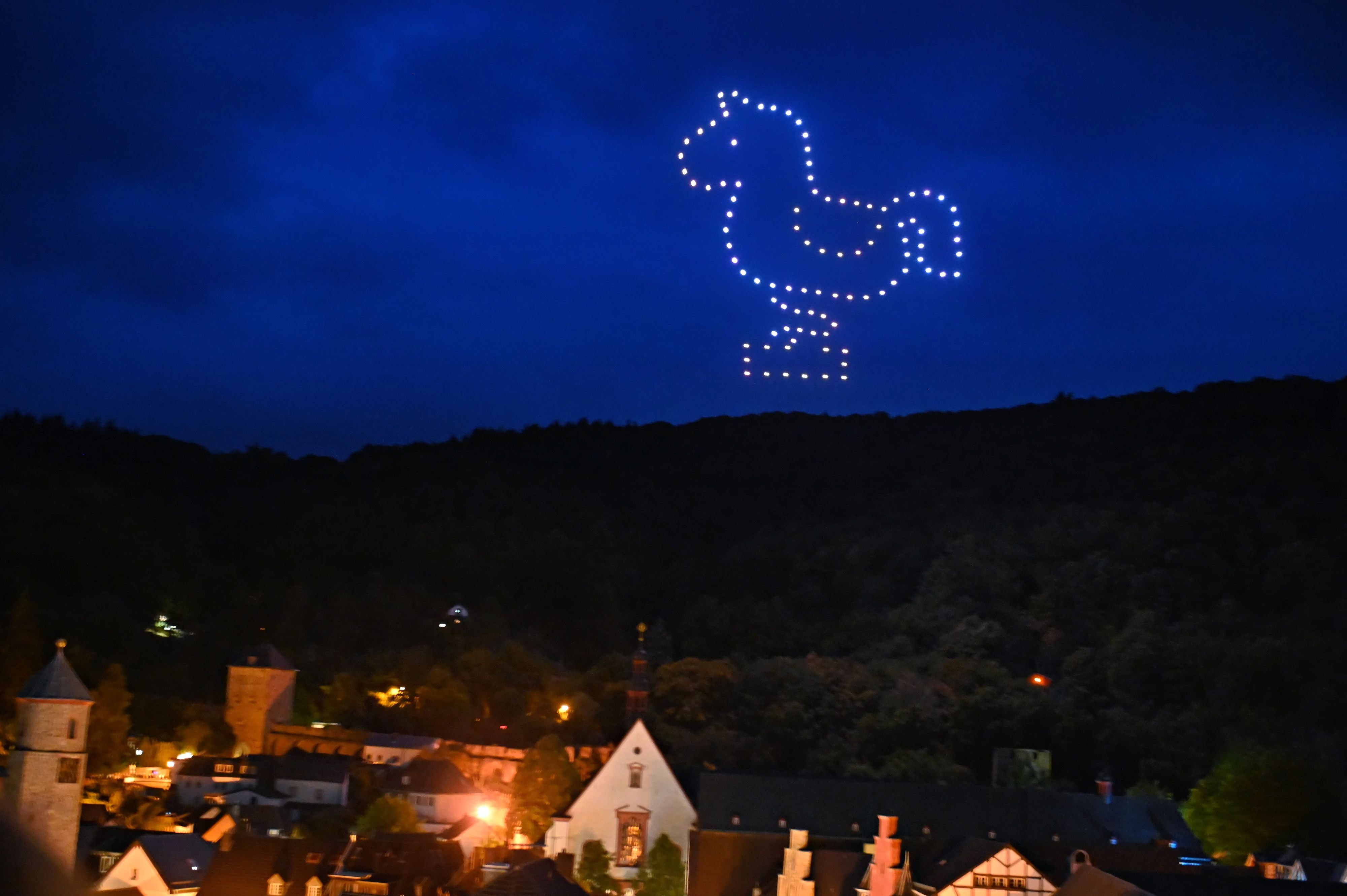 Im tiefblauen Nachhimmel erscheint ein aus leuchtenden Punkten gebildetes Schaukelpferd. Unten am Bildrand sieht man die Häuser der Stadt.