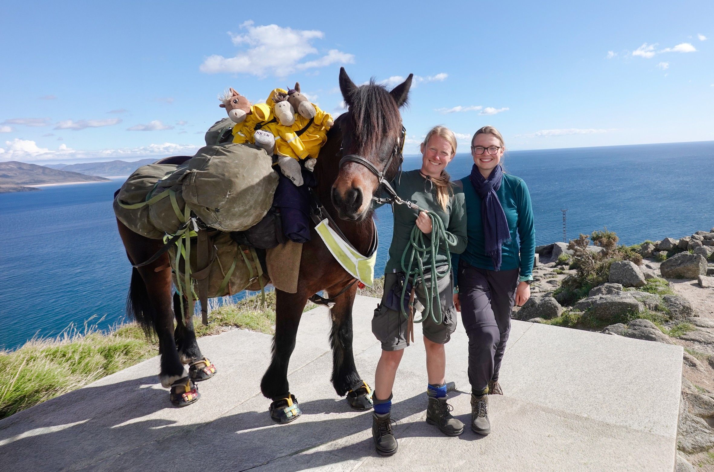 Zu sehen ist ein Pferd mit zwei Frauen. Sie stehen in der Nähe einer Klippe mit dem Rücken zum Meer.
