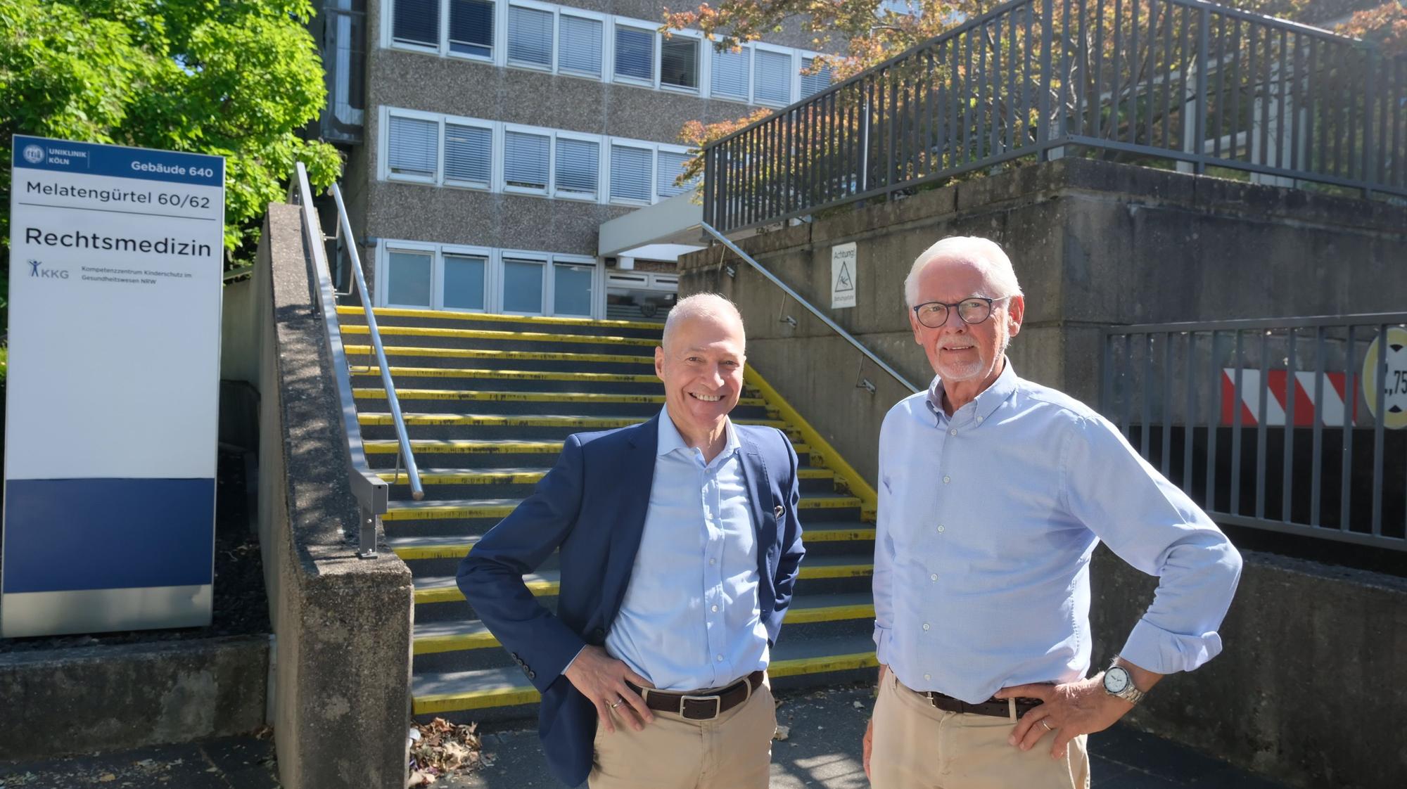 Ein Mann im blauen Blazer und hellblauem Hemd und ein größerer Mann, ebenfalls im hellblauen Hemd, stehen vor der Treppe zum Institut für Rechtsmedizin.
