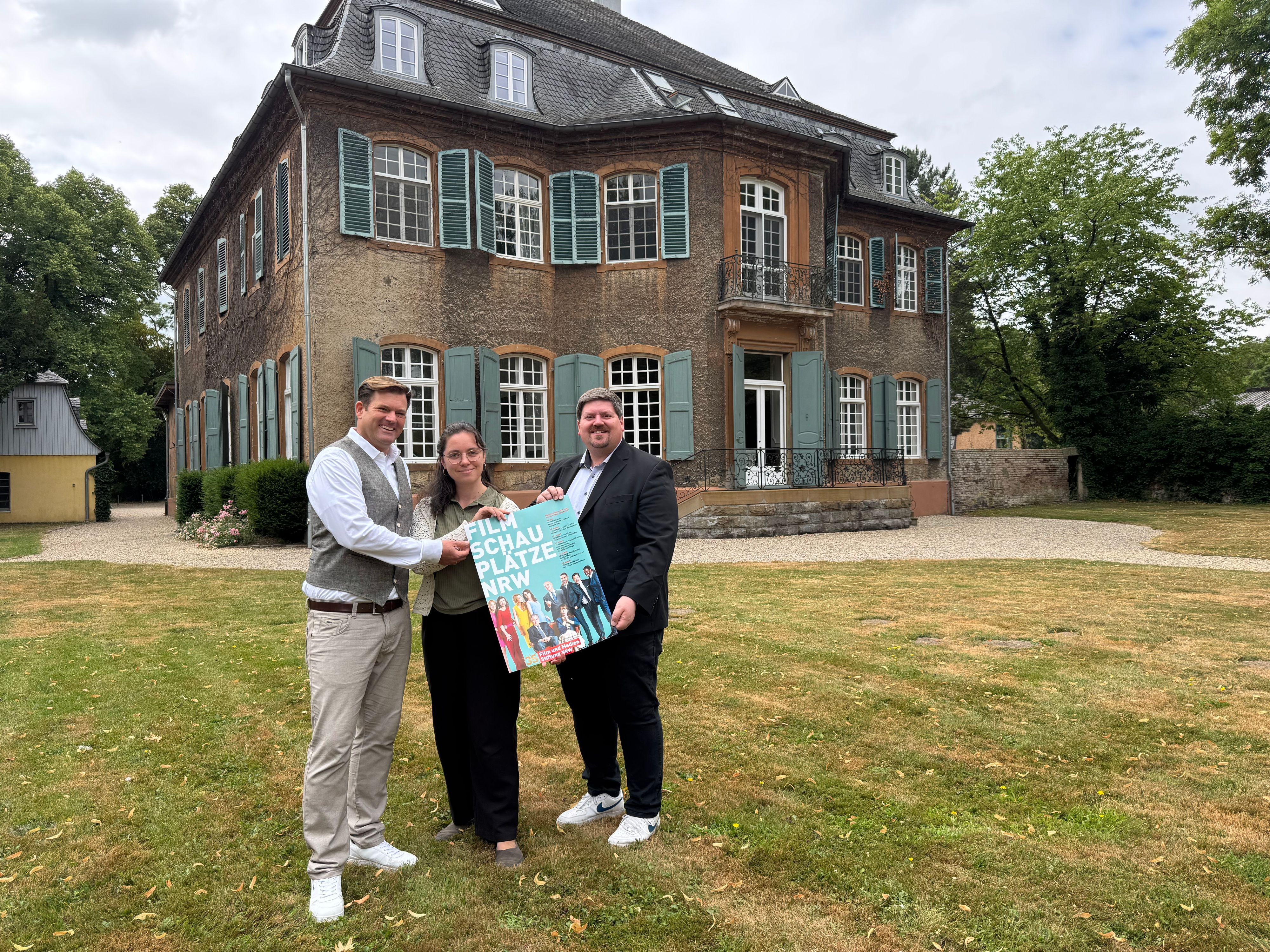 Zwei Männer und eine Frau halten ein Plakat vor einem alten Haus.
