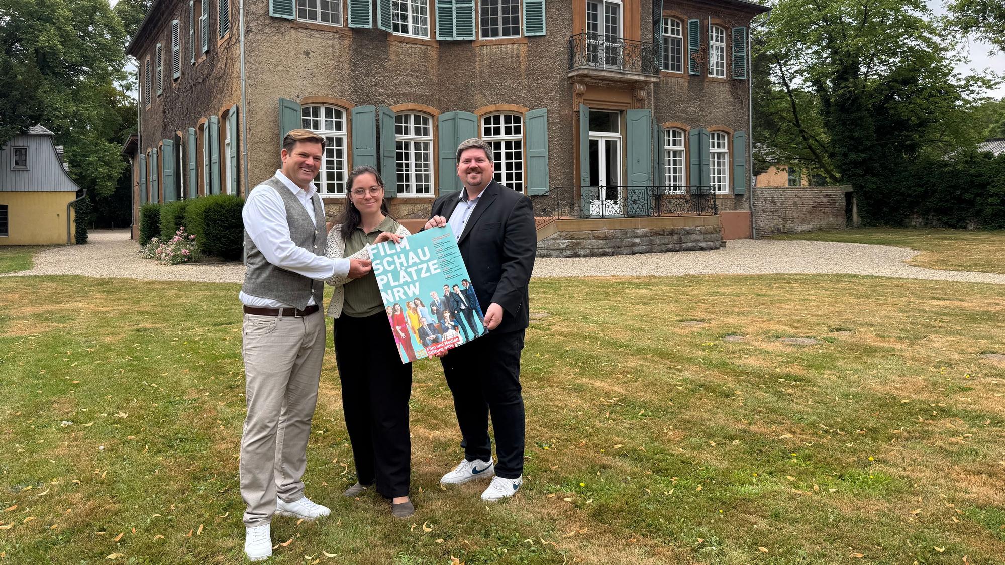Zwei Männer und eine Frau halten ein Plakat vor einem alten Haus.