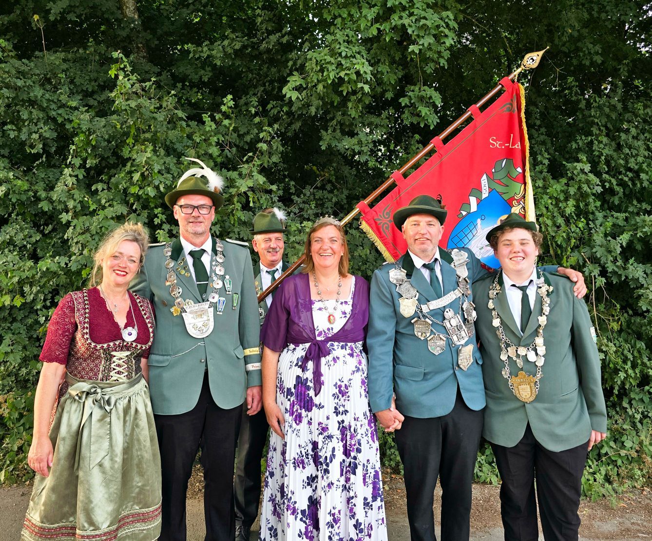 Die neuen Majestäten stehen in einer Reihe, die Frauen in festlichen Kleidern, die Männer in Uniform. Hinter ihnen hält ein Mann eine Standarte hoch.