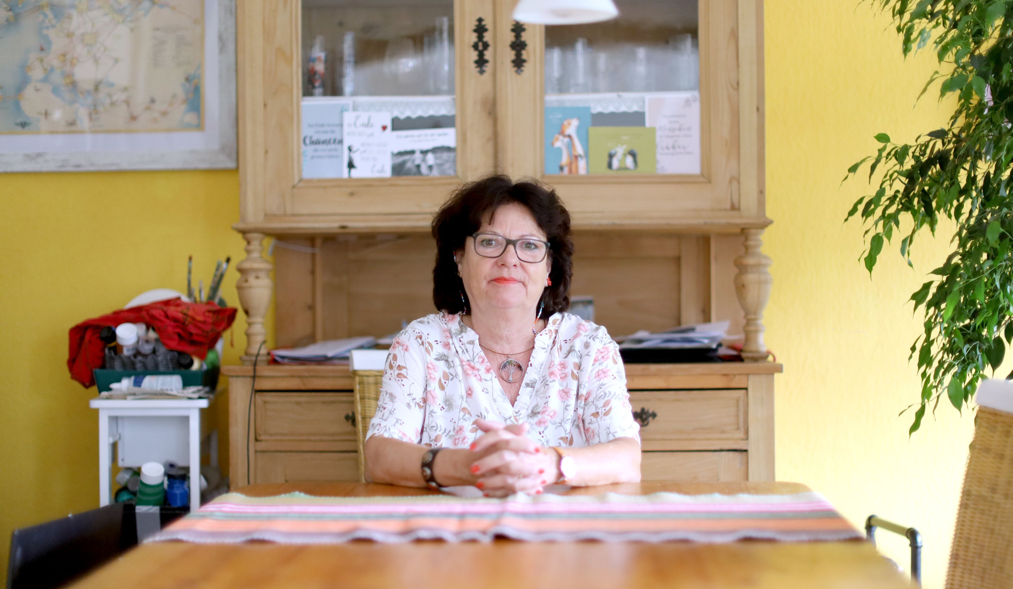 Sylvia Hagen (dunkle Haare, Lippenstift, Bluse mit floralem Print) sitzt an ihrem Küchentisch.