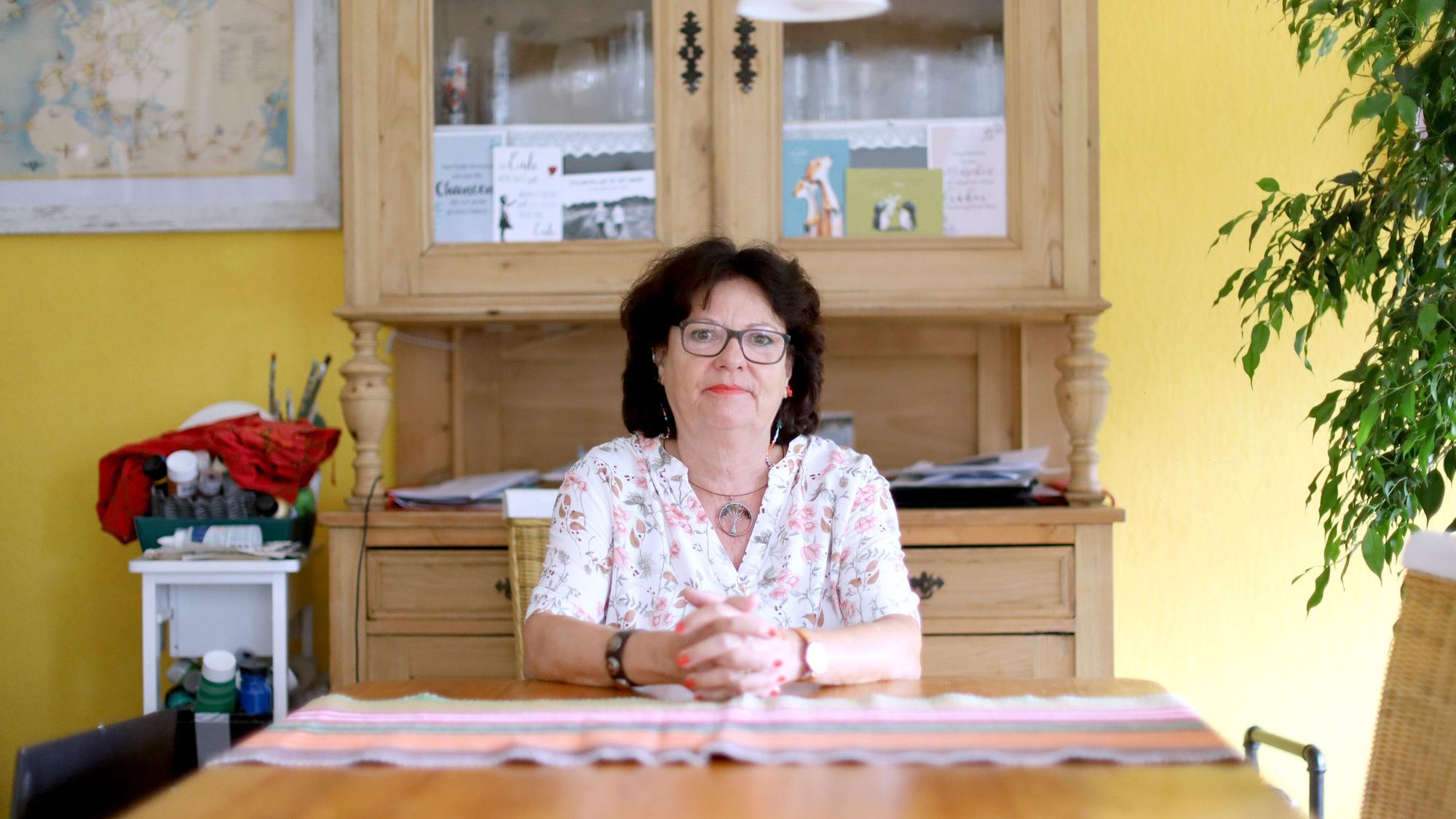 Sylvia Hagen (dunkle Haare, Lippenstift, Bluse mit floralem Print) sitzt an ihrem Küchentisch.