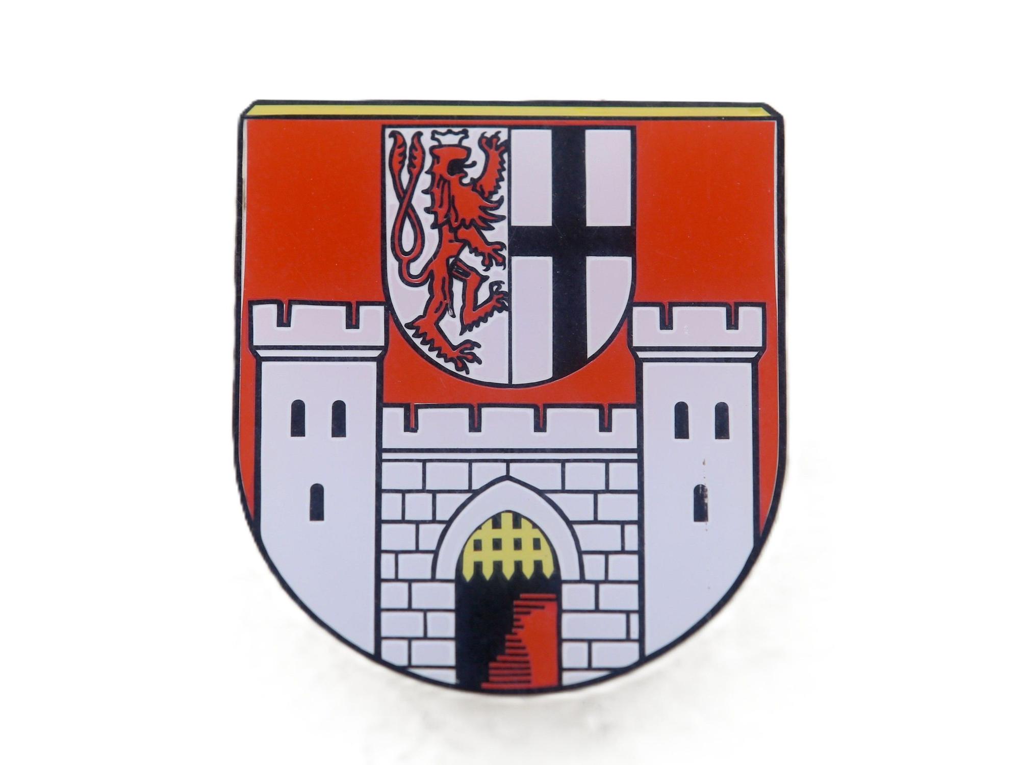 Das Wappen der Stadt Königswinter enthält eine Burg mit einem offenen Fallgatter und darüber ein Schild in dem links ein Bergischer Löwe in Richtung des Kurfürstenkreuzes nach rechts schaut.