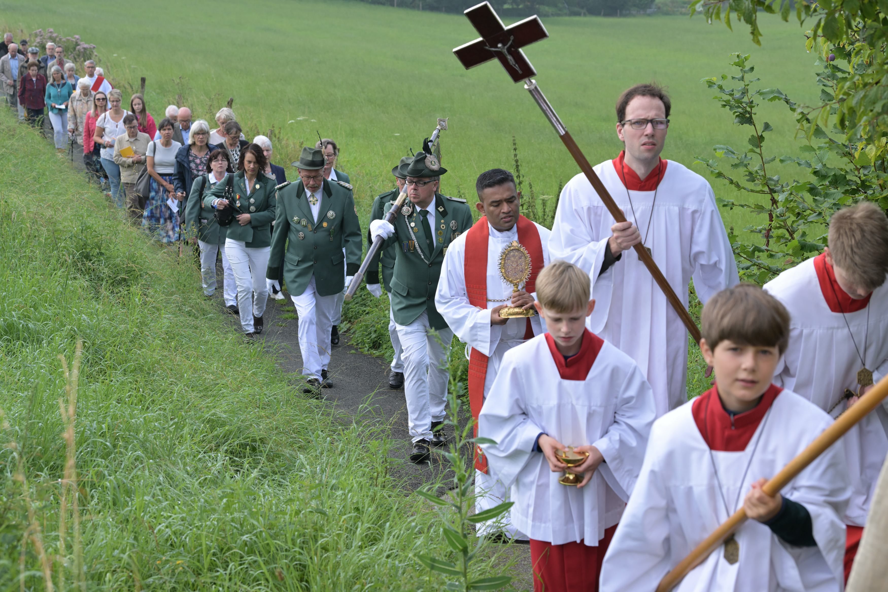 Zu sehen sind viele Menschen, die in einer Schlange einen Weg entlang laufen. Im Vordergrund ist ein Geistlicher mit einem großen Kreuz an einem Stab zu sehen.