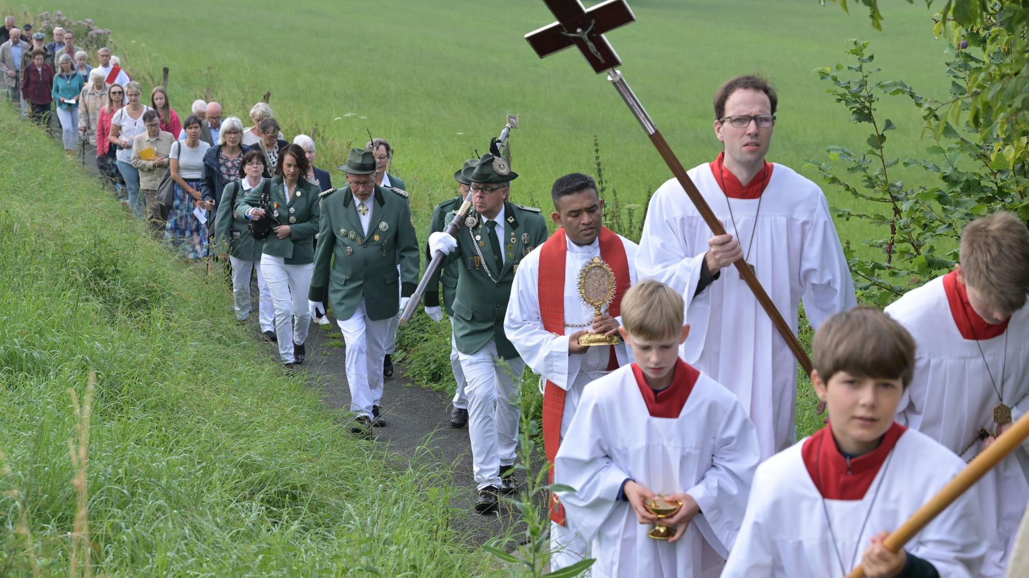 Zu sehen sind viele Menschen, die in einer Schlange einen Weg entlang laufen. Im Vordergrund ist ein Geistlicher mit einem großen Kreuz an einem Stab zu sehen.