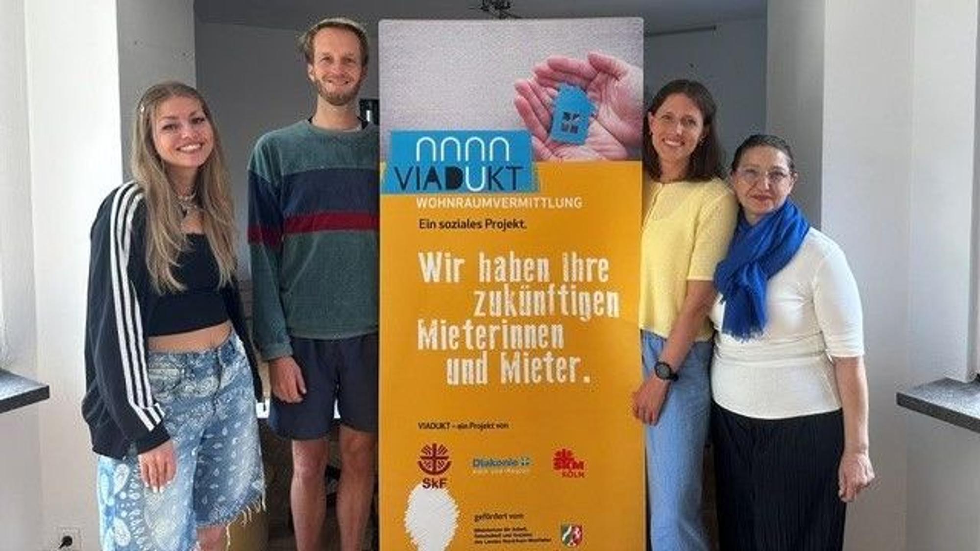 Drei Frauen und ein Mann stehen links und rechts von einem Aufsteller, auf dem steht „Wir haben Ihre zukünftigen Mieterinnen und Mieter“.