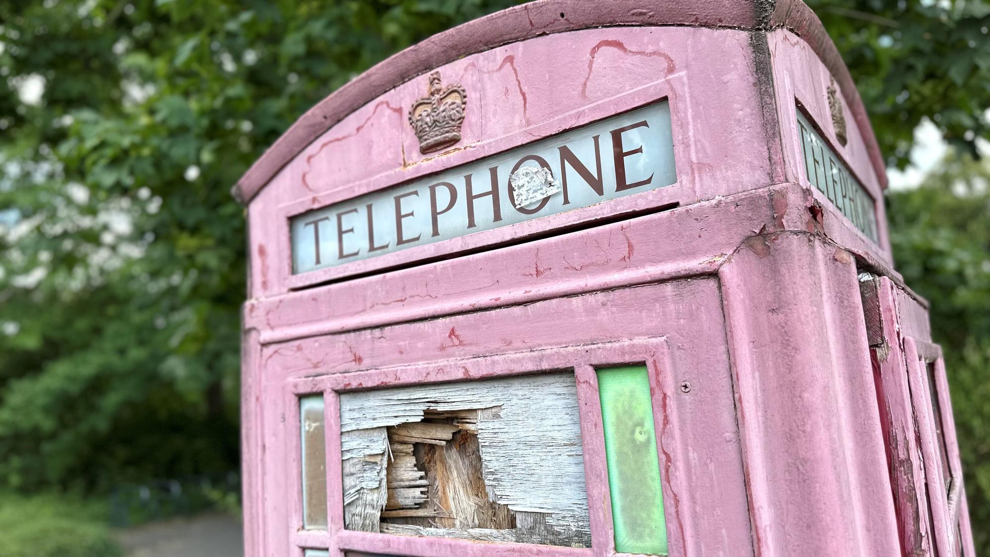 Eine rote Telefonzelle steht auf einem Platz, dahinter sind Beete und Bäume zu sehen.