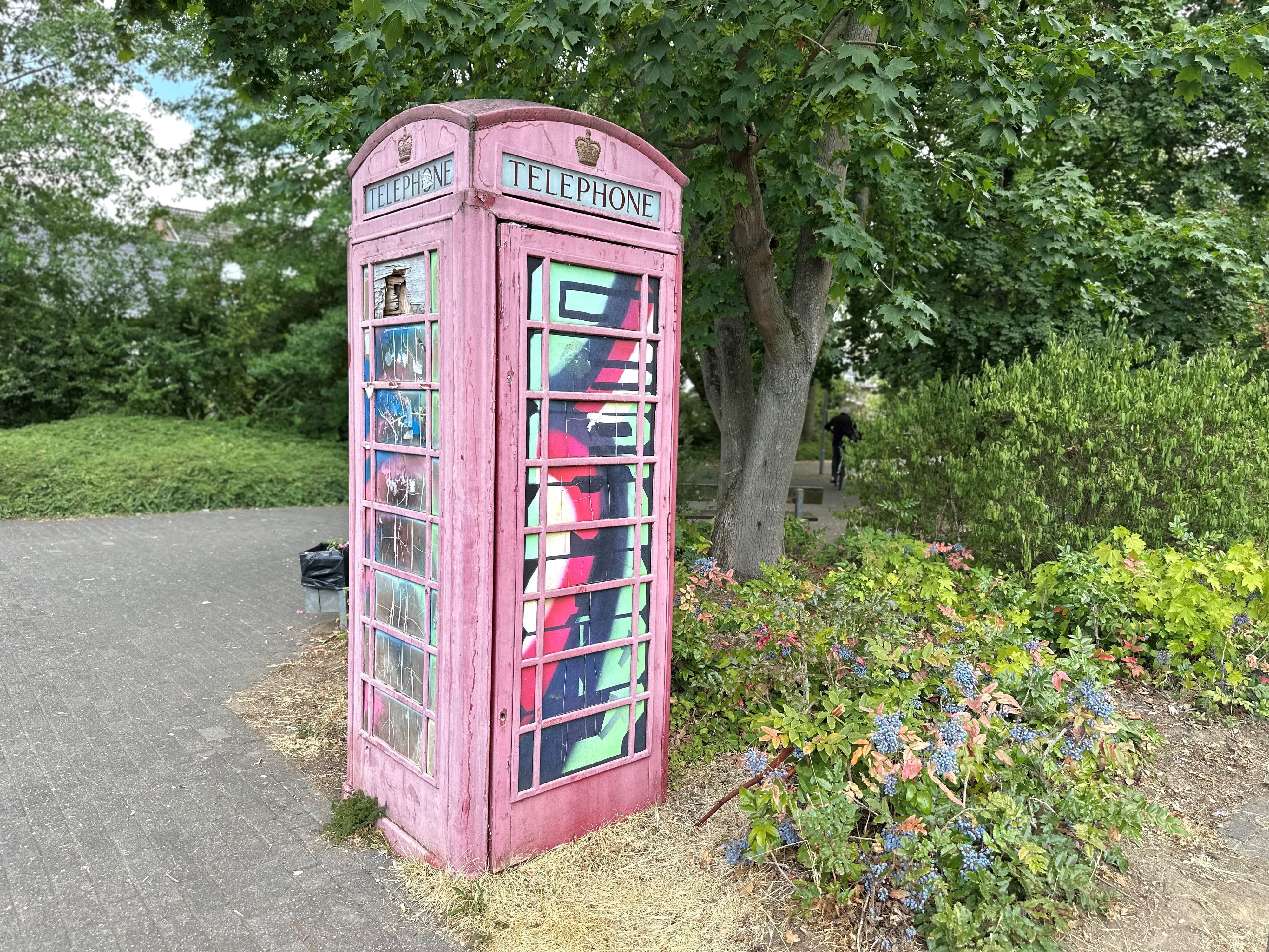 Eine rote Telefonzelle steht auf einem Platz, dahinter sind Beete und Bäume zu sehen.