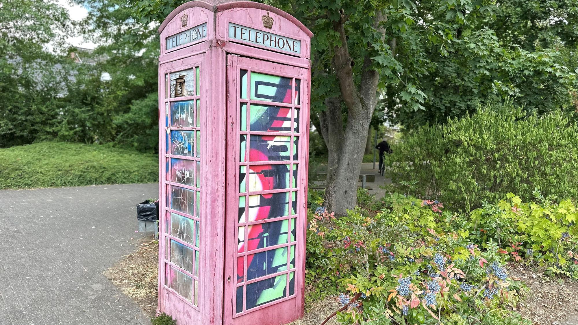 Eine rote Telefonzelle steht auf einem Platz, dahinter sind Beete und Bäume zu sehen.