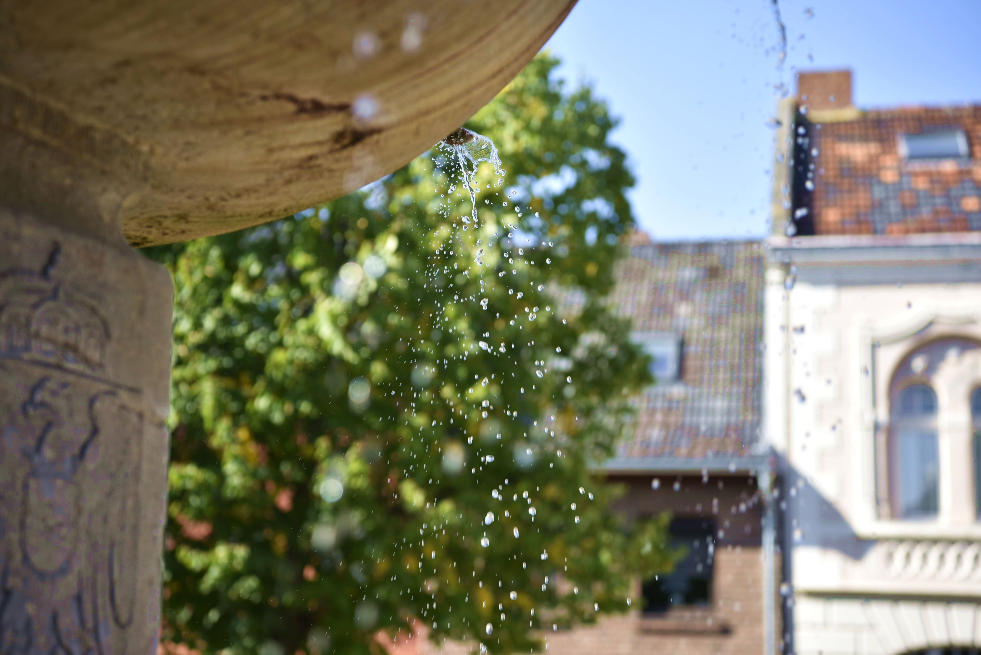 Aus einem Brunnen tropft Wasser, im Hintergrund sind Häuser und Bäume zu erkennen.