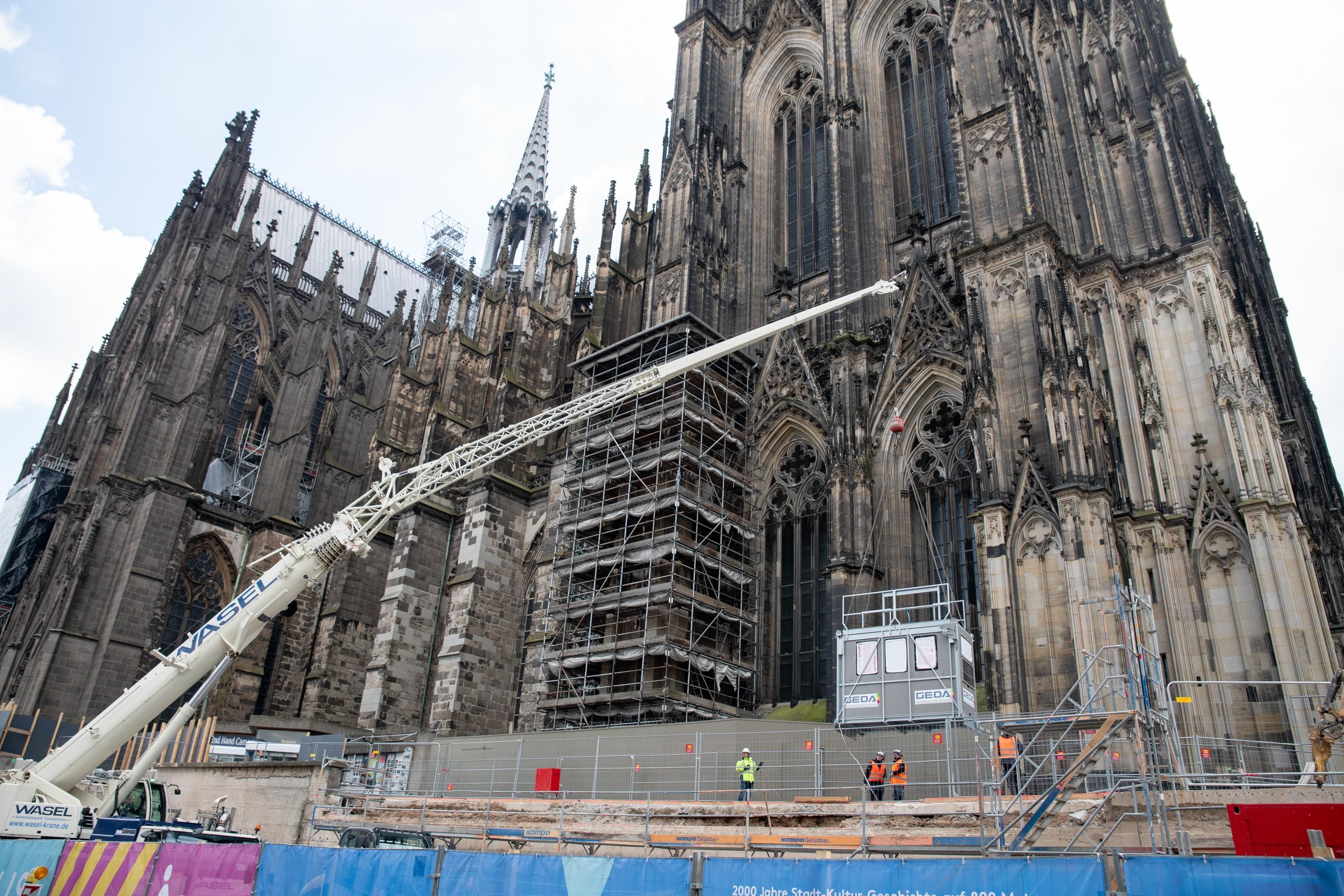 Vor dem Kölner Dom hängt eine Aufzugkabine am Haken eines Mobilkrans.