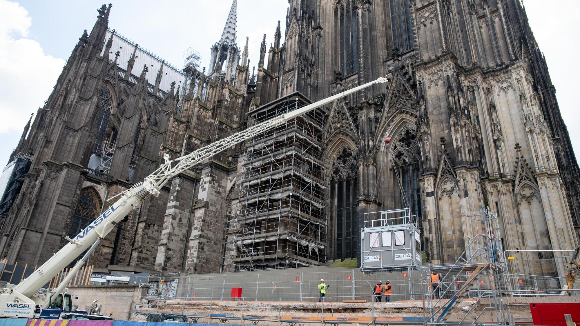 Vor dem Kölner Dom hängt eine Aufzugkabine am Haken eines Mobilkrans.