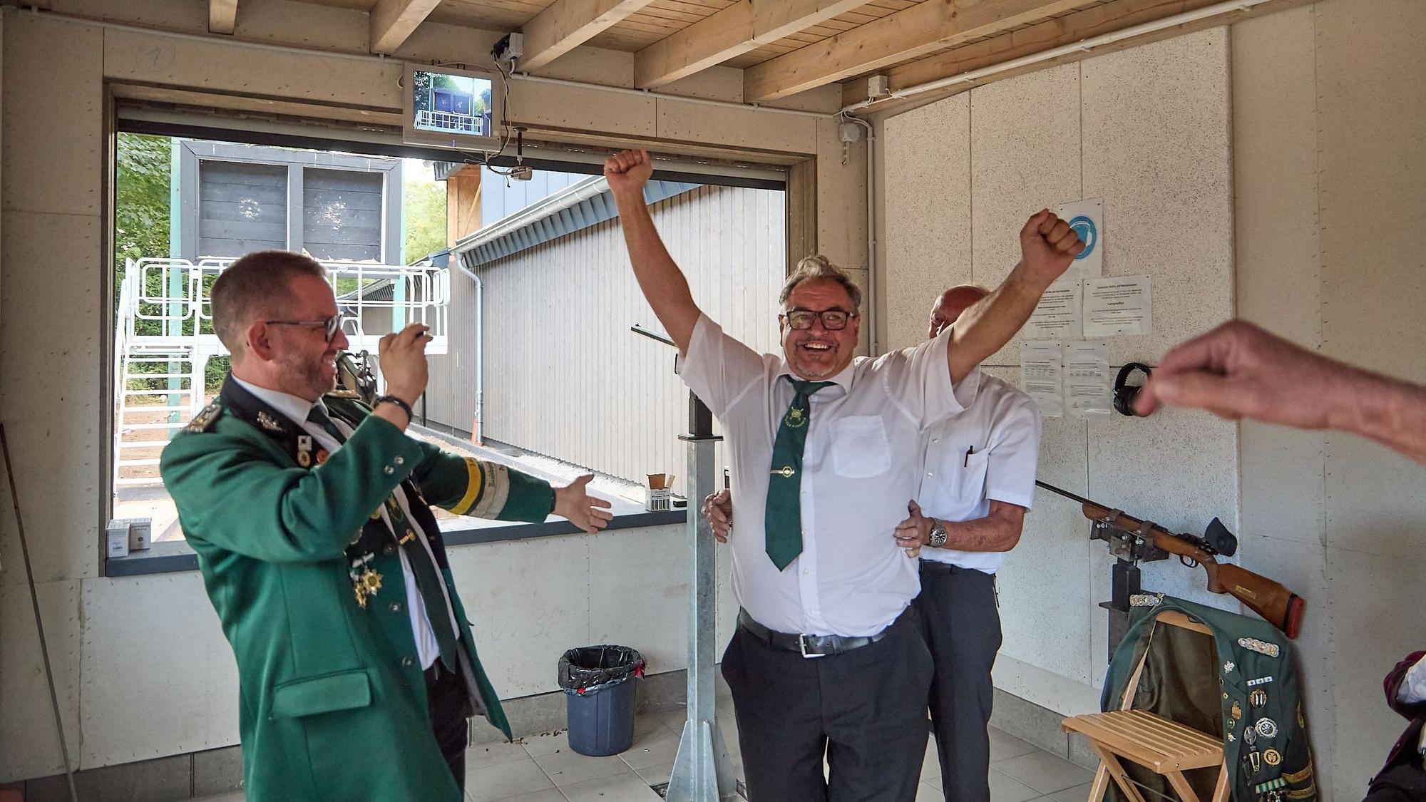 Ein Mann in weißem Hemd und mit grüner Krawatte reckt beide Arme in die Luft und freut sich. Neben ihm steht ein Mann in grüner Schützenuniform und lacht ihn an.
